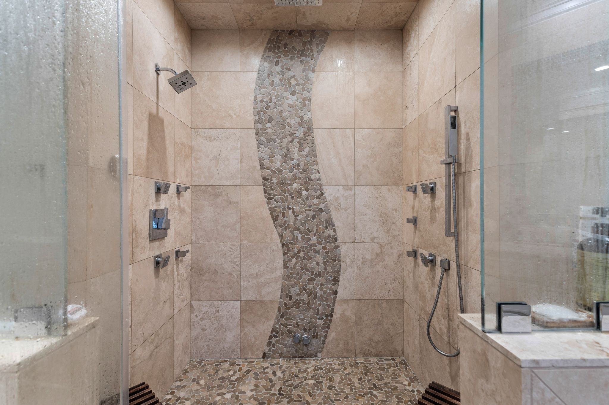 Walk-in shower with beige stone tile, a curved pebble-mosaic accent, and multiple shower fixtures.