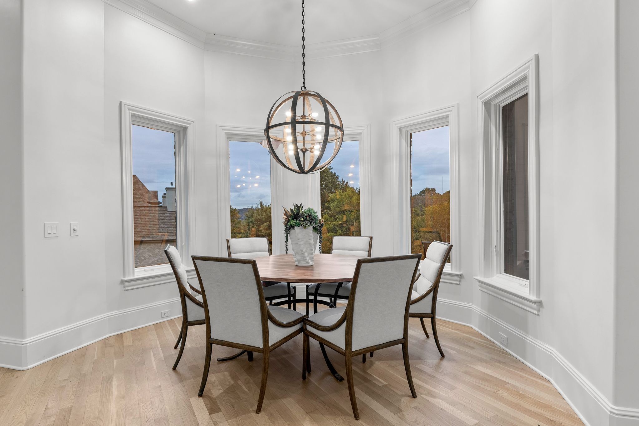 Dining room with a round table, spherical chandelier, and tall windows overlooking autumn trees.