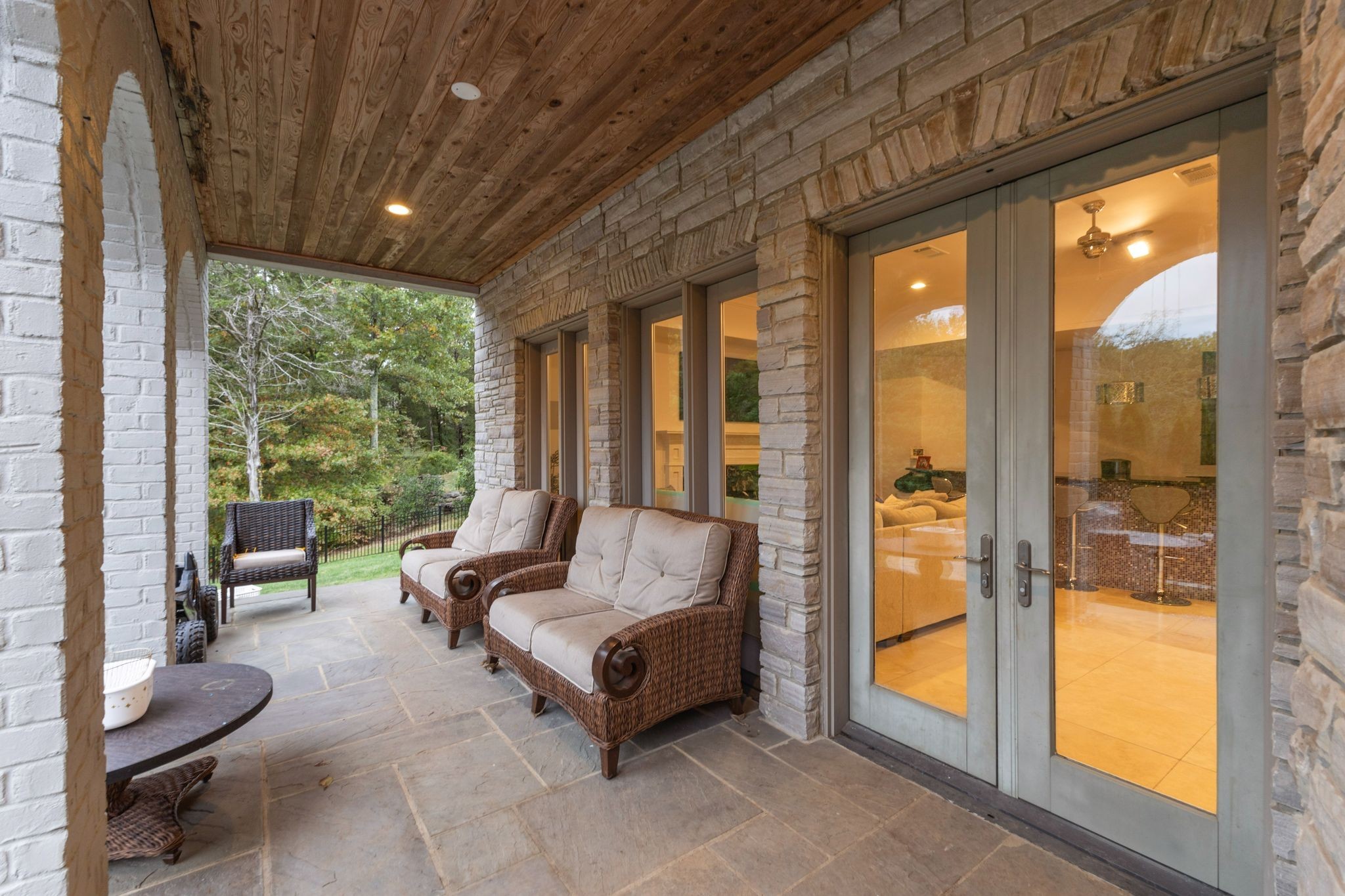 Covered stone patio with wicker seating, a wood ceiling, and glass doors opening to the home.