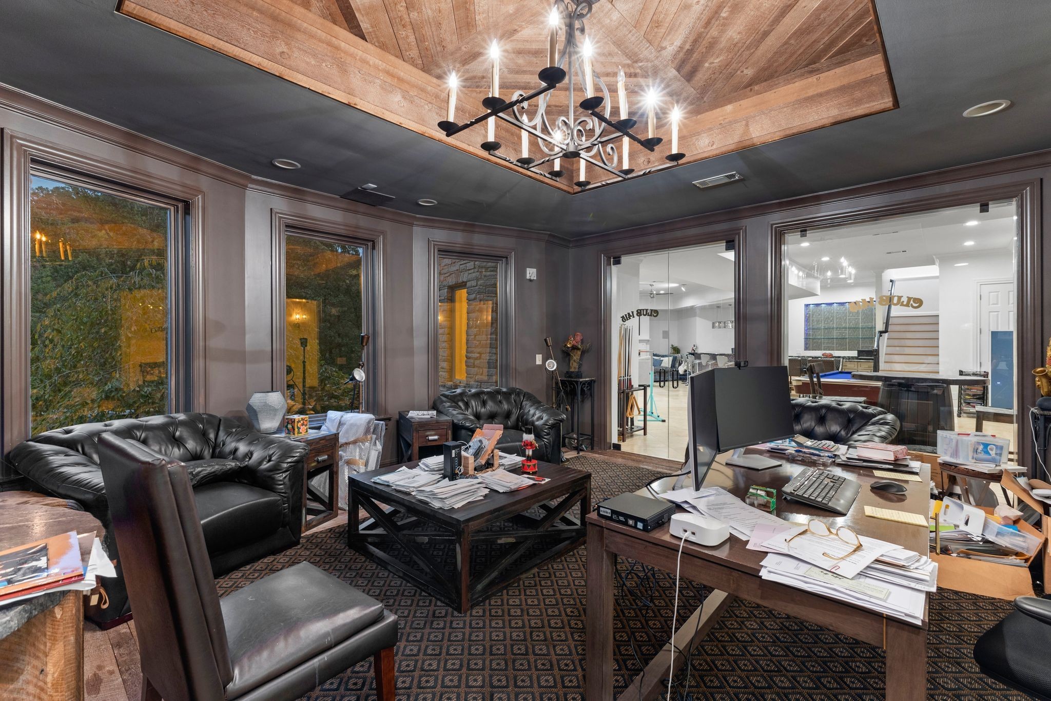 Office with leather seating, paper-covered desk, and recessed wood ceiling beside a room with a pool table.