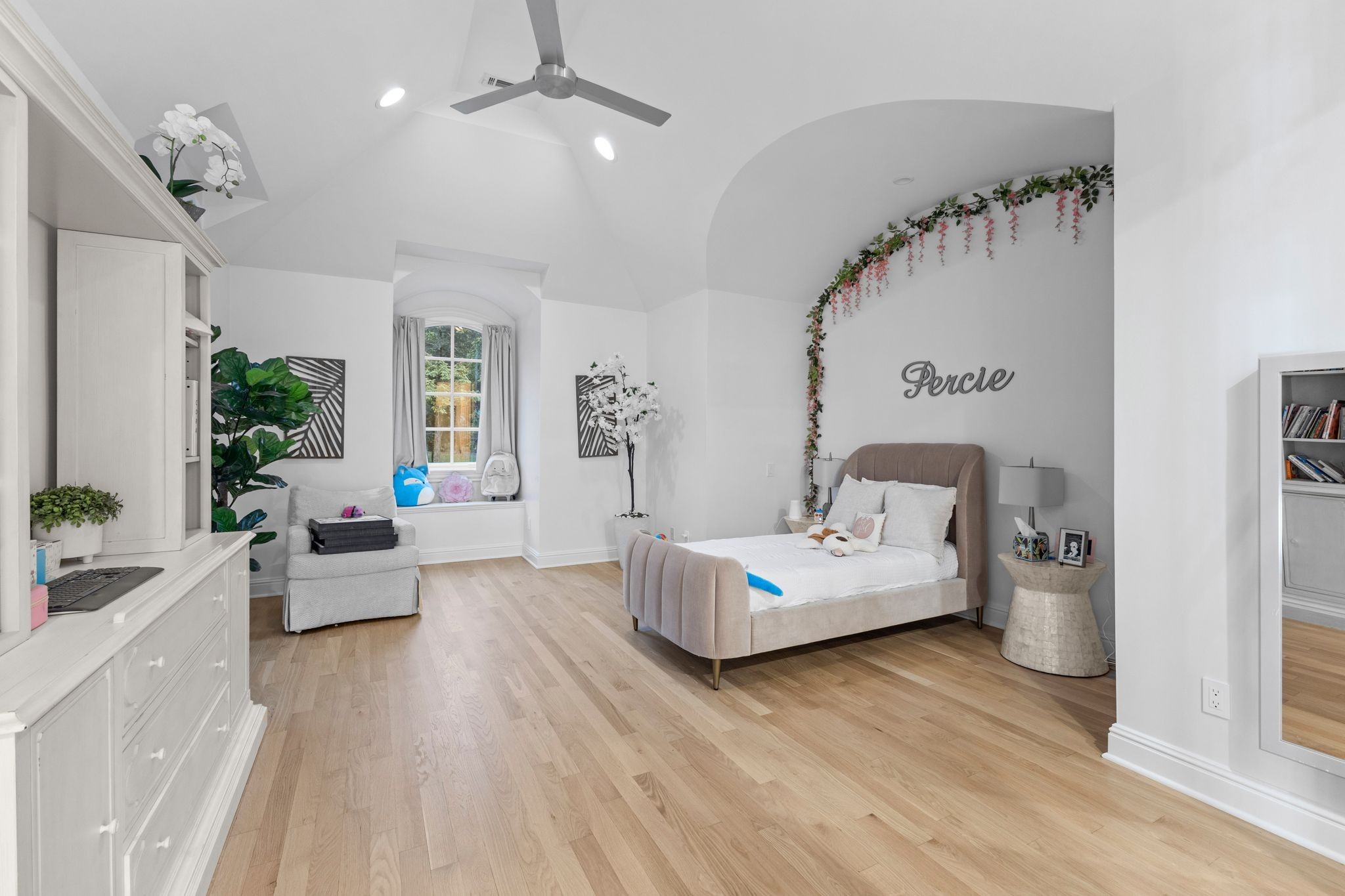 Furnished bedroom with a vaulted ceiling, arched window, and upholstered bed beneath a floral garland.