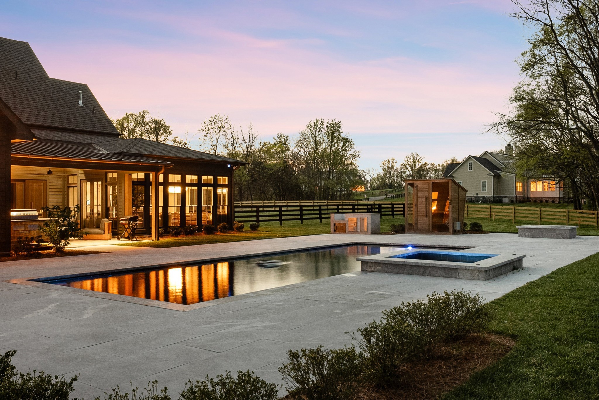 Backyard pool and raised spa beside a broad patio and warmly lit covered porch at dusk.