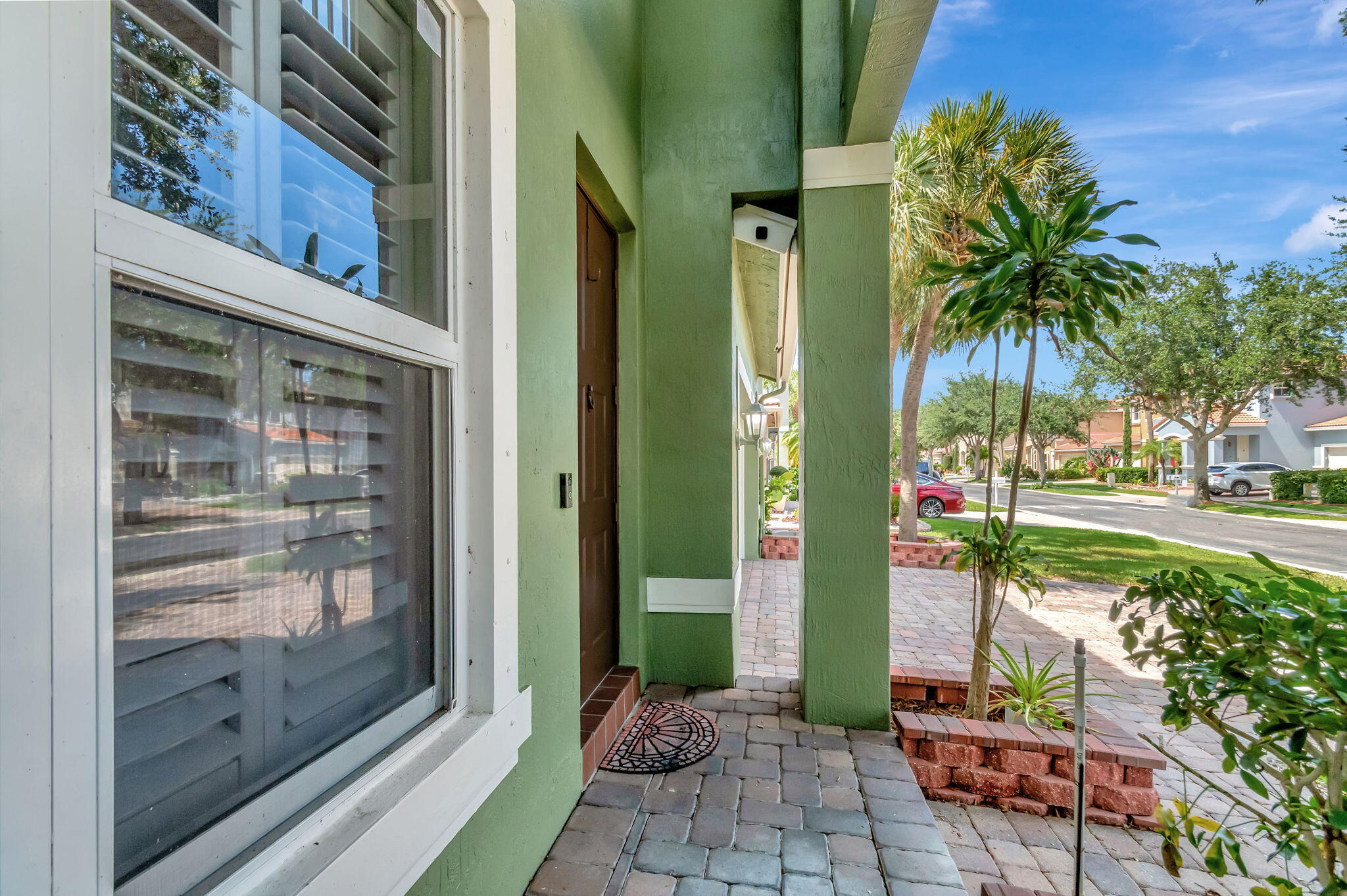Green home’s covered front entry with a brown door, paver walkway, and tropical landscaping.