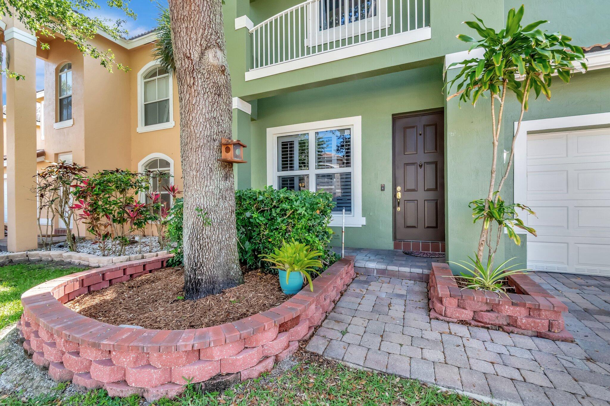 Green two-story home entrance with a paved walkway, raised brick planters, and a large shade tree.