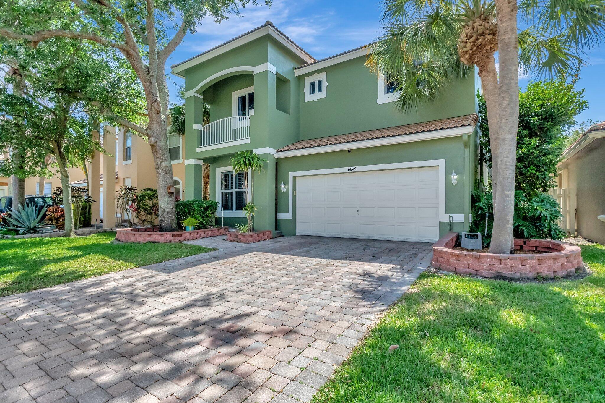 Green two-story house with a white double garage, paver driveway, and mature palm trees.
