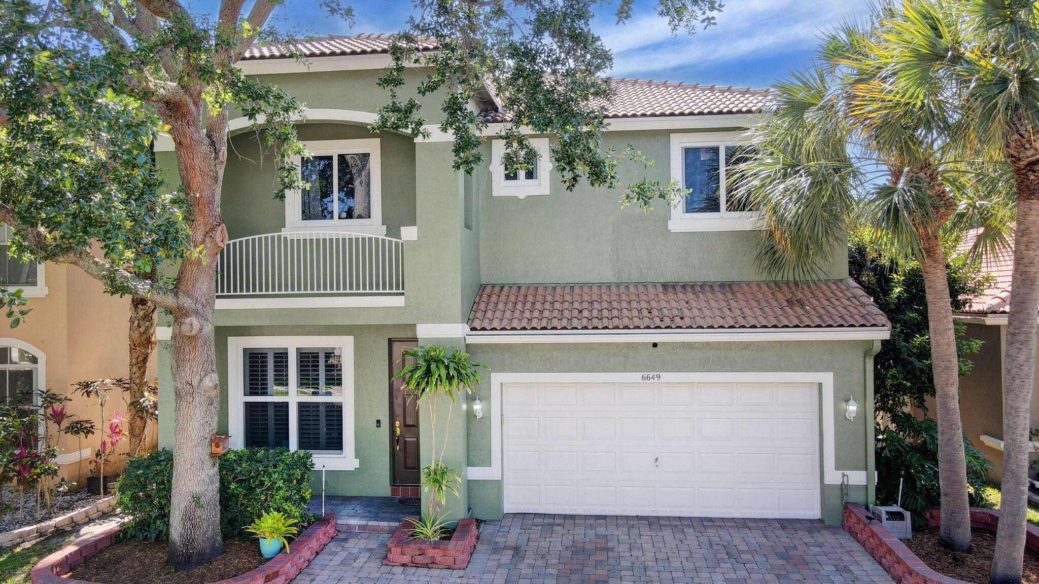 Two-story sage-green house with a white double garage door, tiled roof, and palm trees.