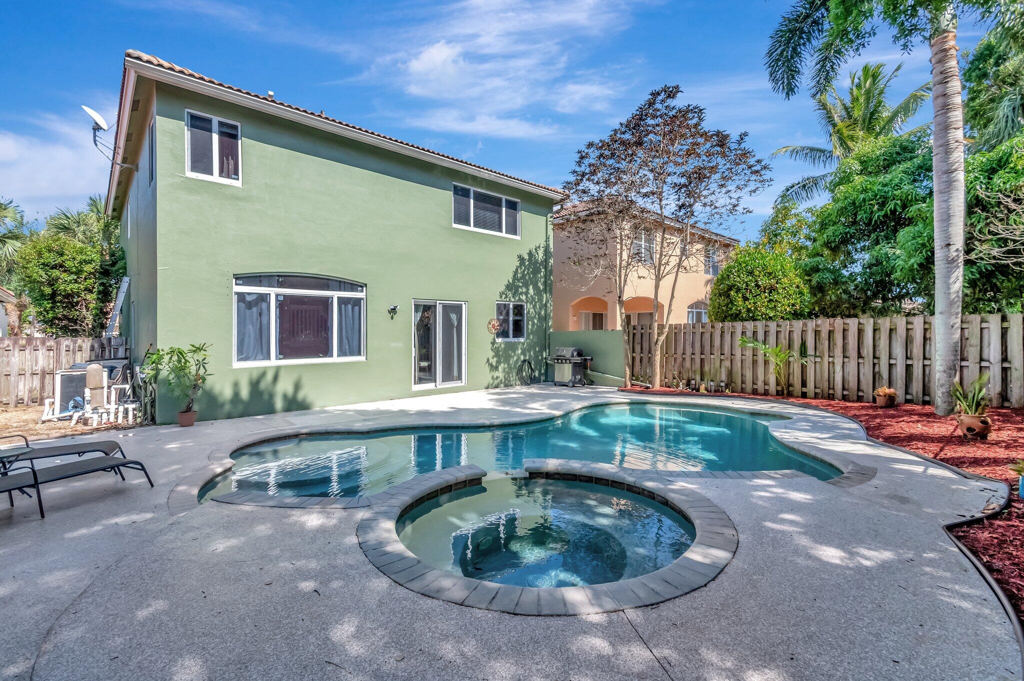 Backyard pool and circular spa beside a two-story green house and fenced tropical garden.
