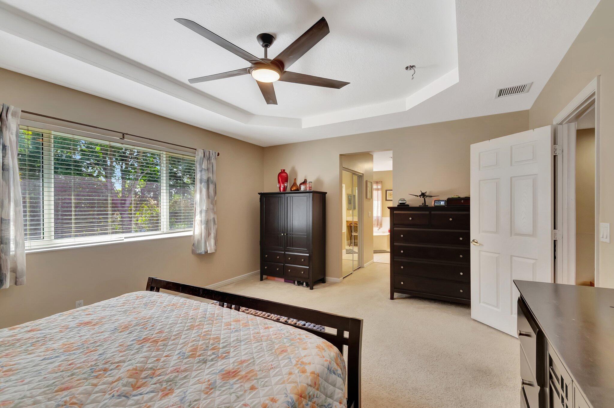 Bedroom with a tray ceiling, dark ceiling fan, large window, and dark wood dressers.