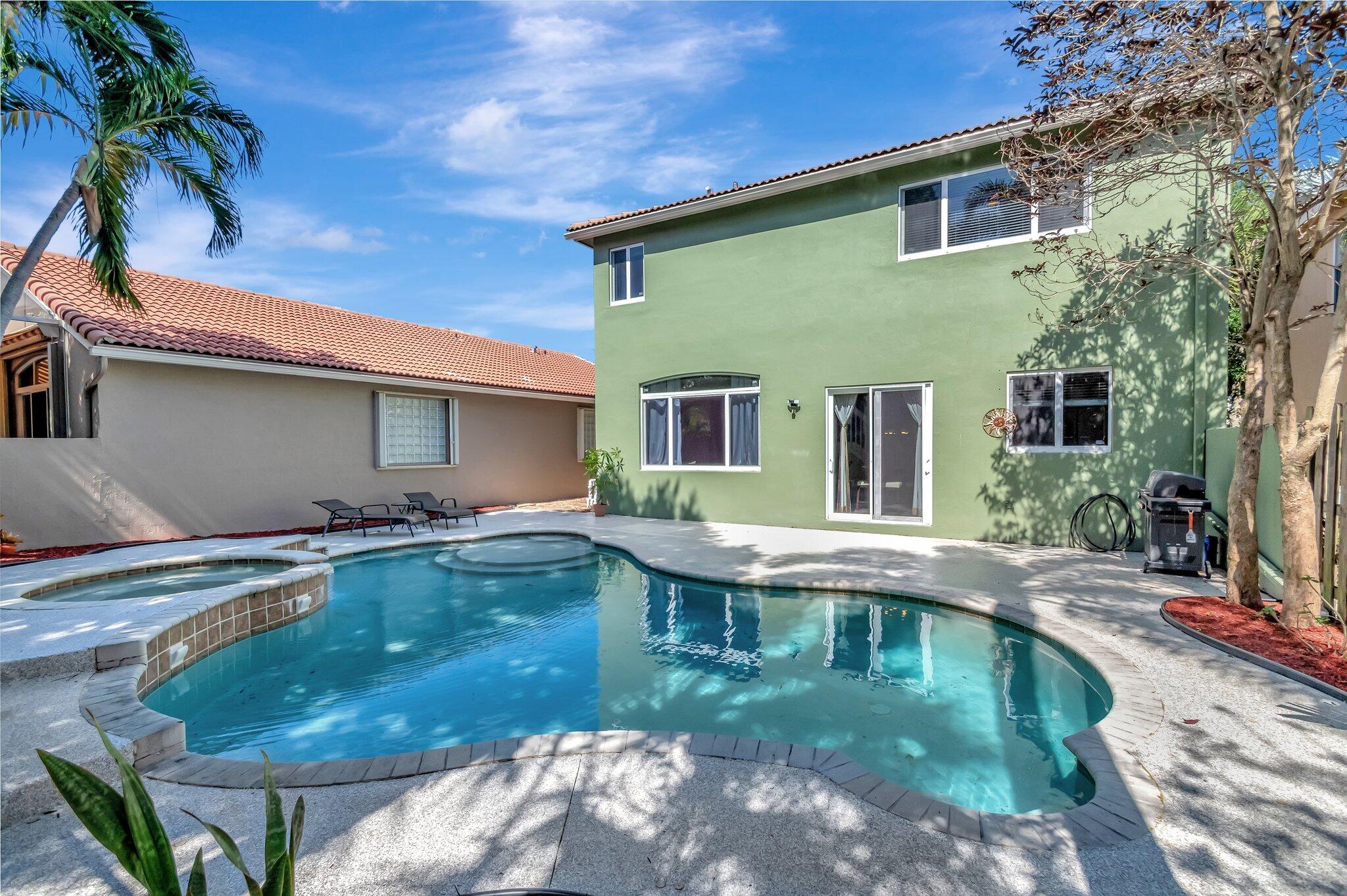 Backyard pool and spa beside a two-story green stucco home, with palm trees and patio seating.