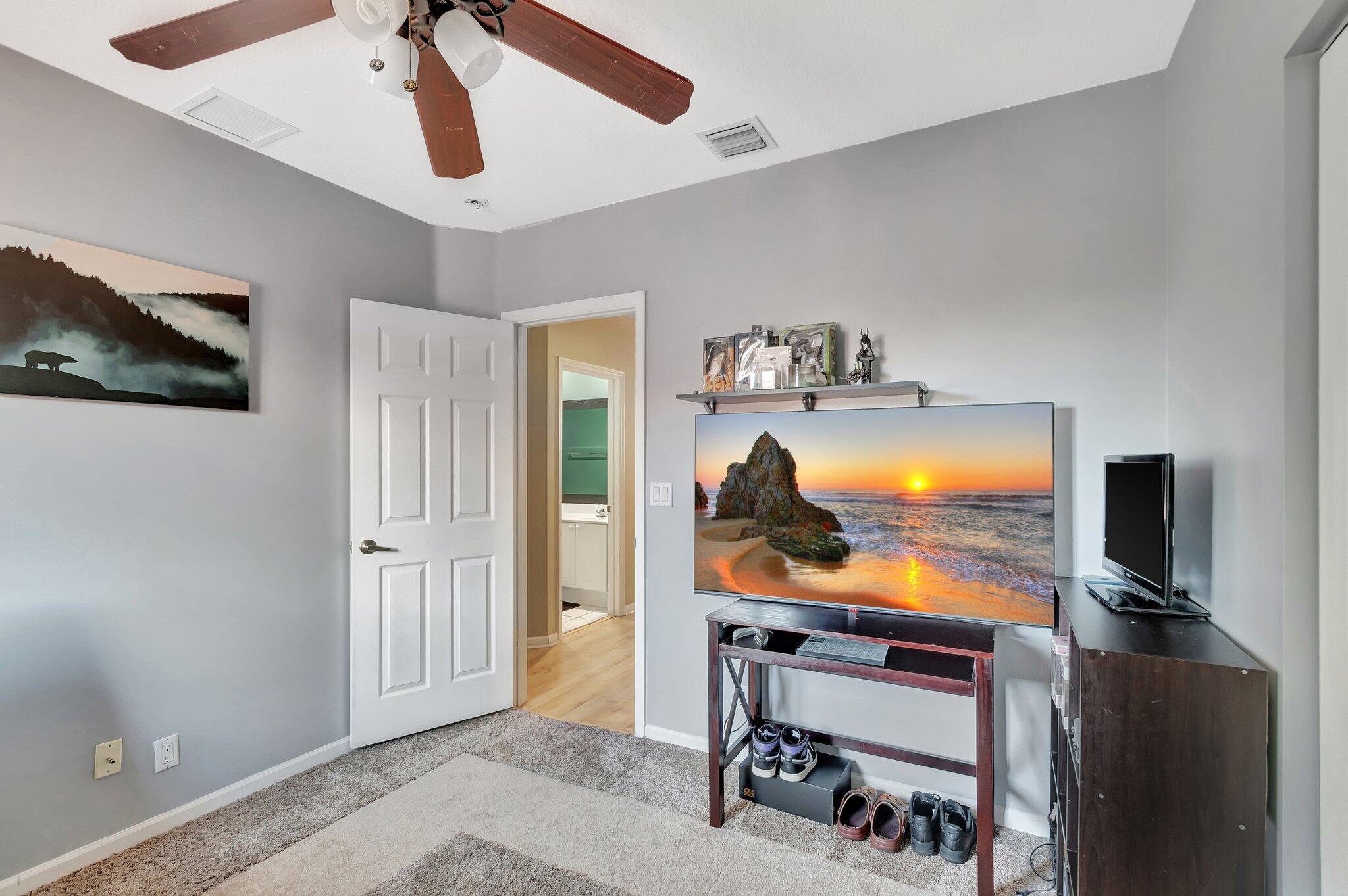 Furnished bedroom with a large television, ceiling fan, and open doorway to the bathroom.