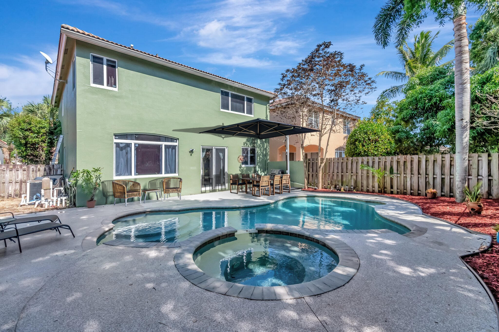 Backyard pool and circular spa beside a green two-story house, with a shaded outdoor dining area.
