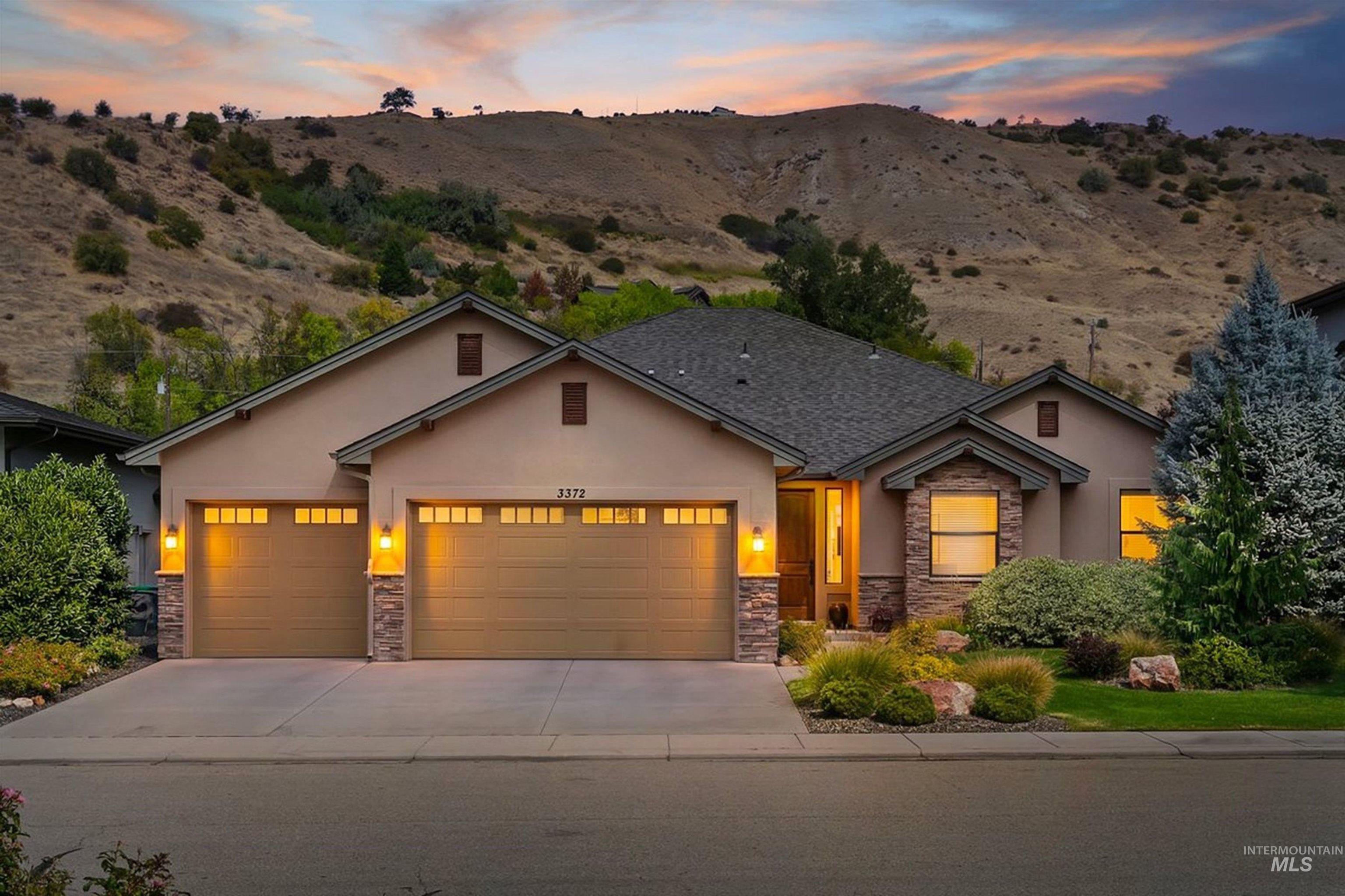 Beige stucco home with stone accents and a three-car garage, glowing at dusk beneath dry foothills.