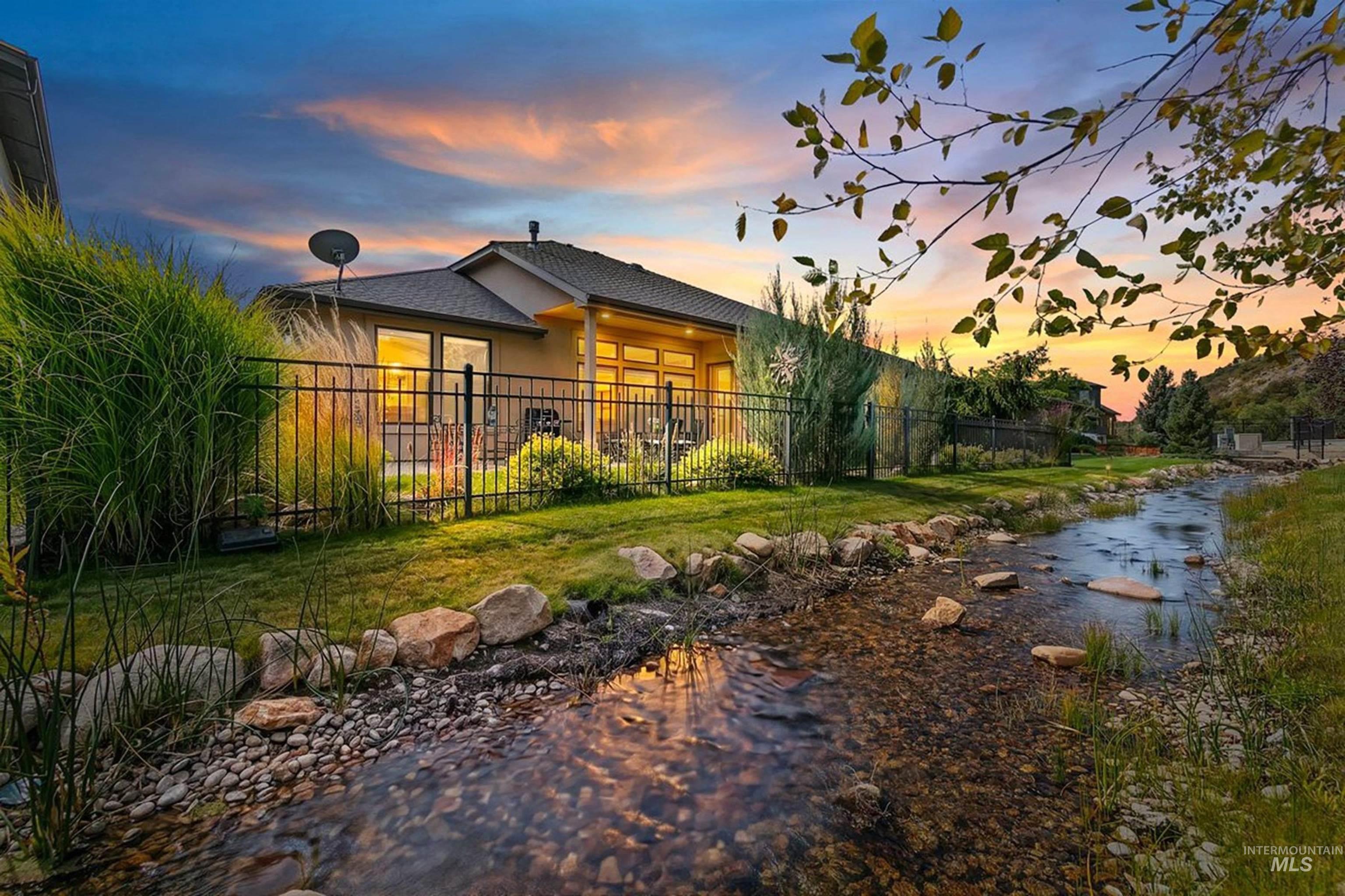 Home beside a shallow rock-lined creek, with a fenced yard glowing at sunset.