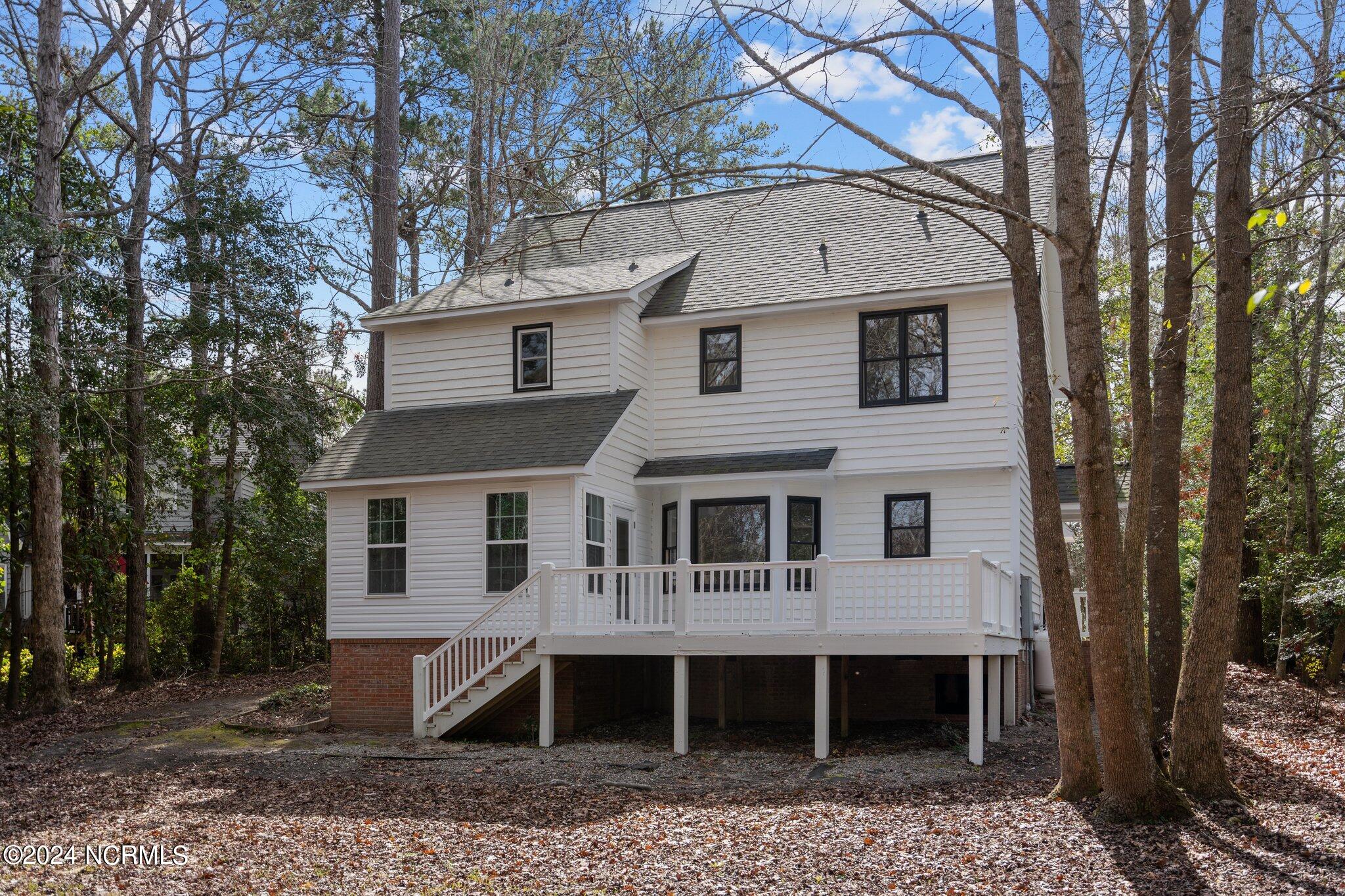 Rear of a two-story white house with a raised deck and stairs, surrounded by mature trees.