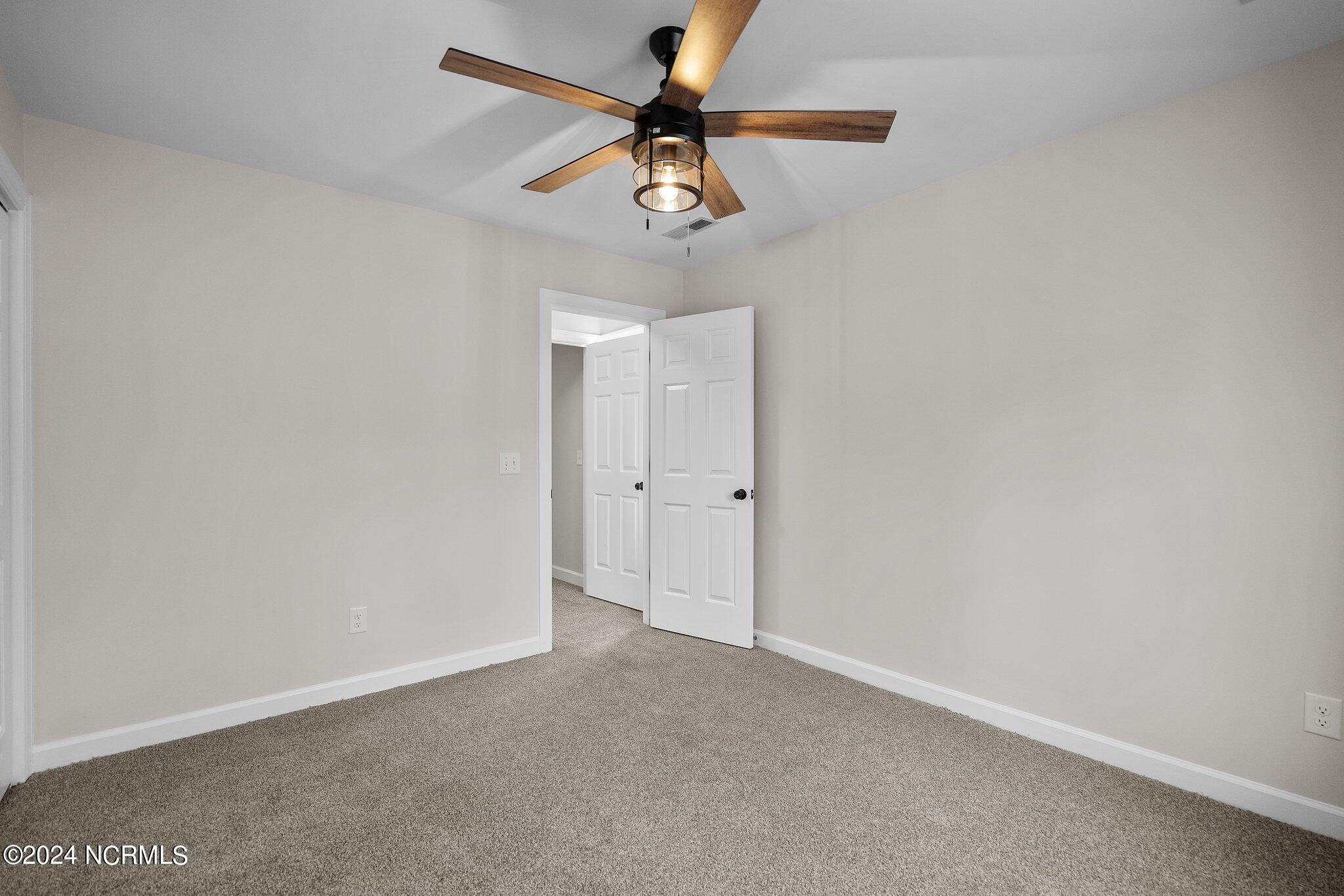 Empty bedroom with gray carpet, an illuminated wood-blade ceiling fan, and an open white door.