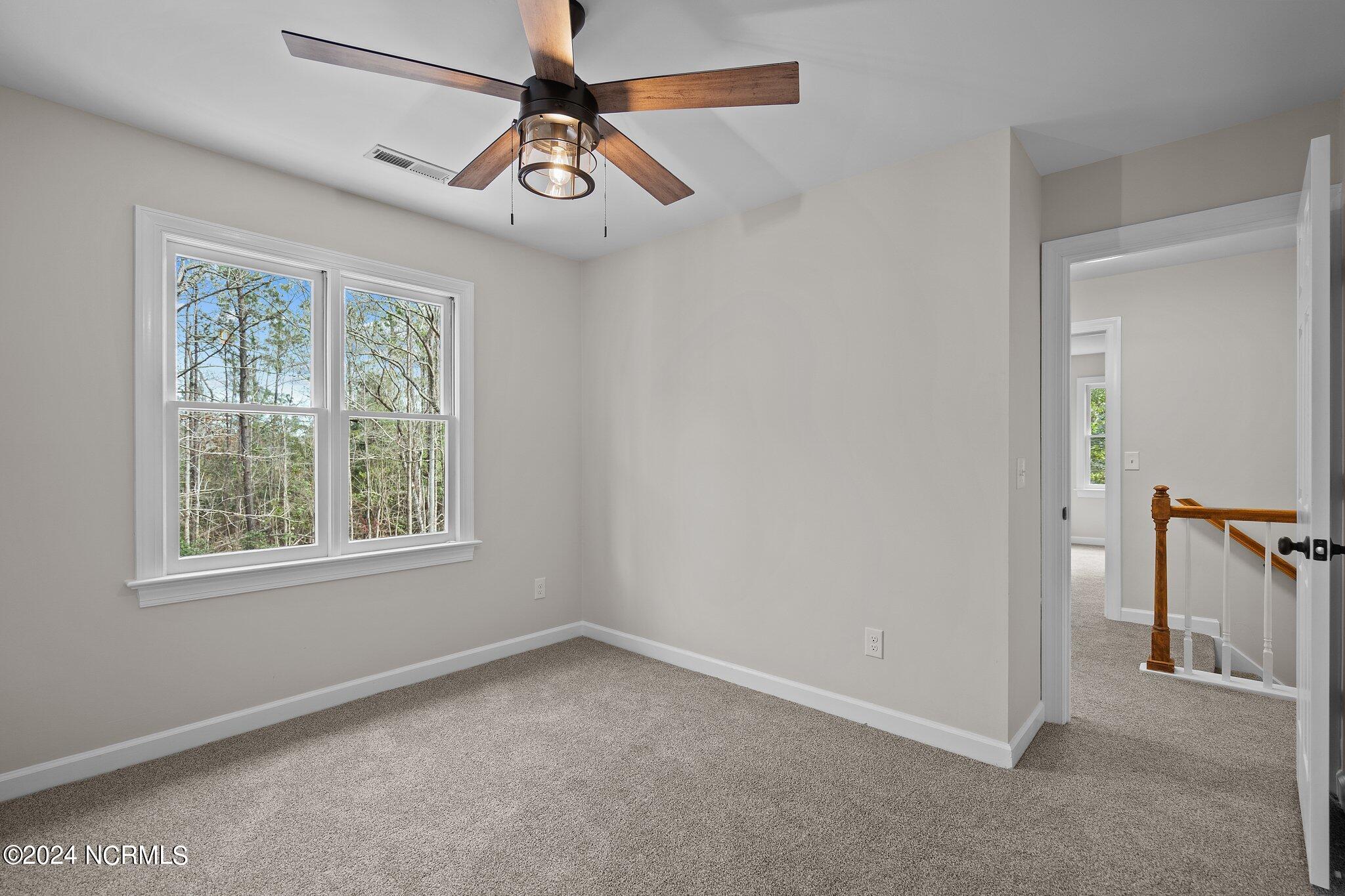 Empty carpeted bedroom with two tree-facing windows, a wood-blade ceiling fan, and an open doorway to the landing.