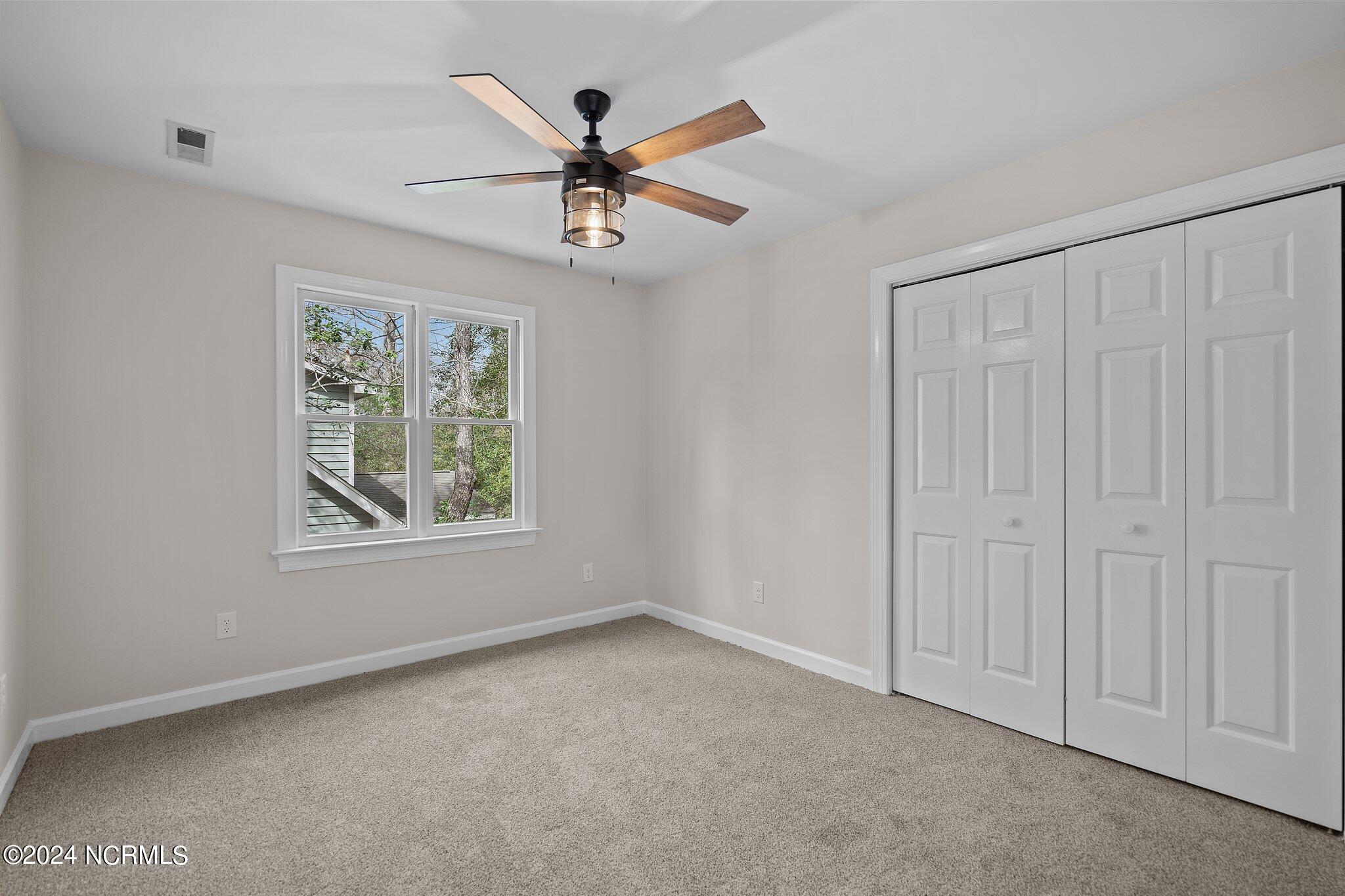 Empty bedroom with beige carpet, a double window, white closet doors, and a wood-blade ceiling fan.