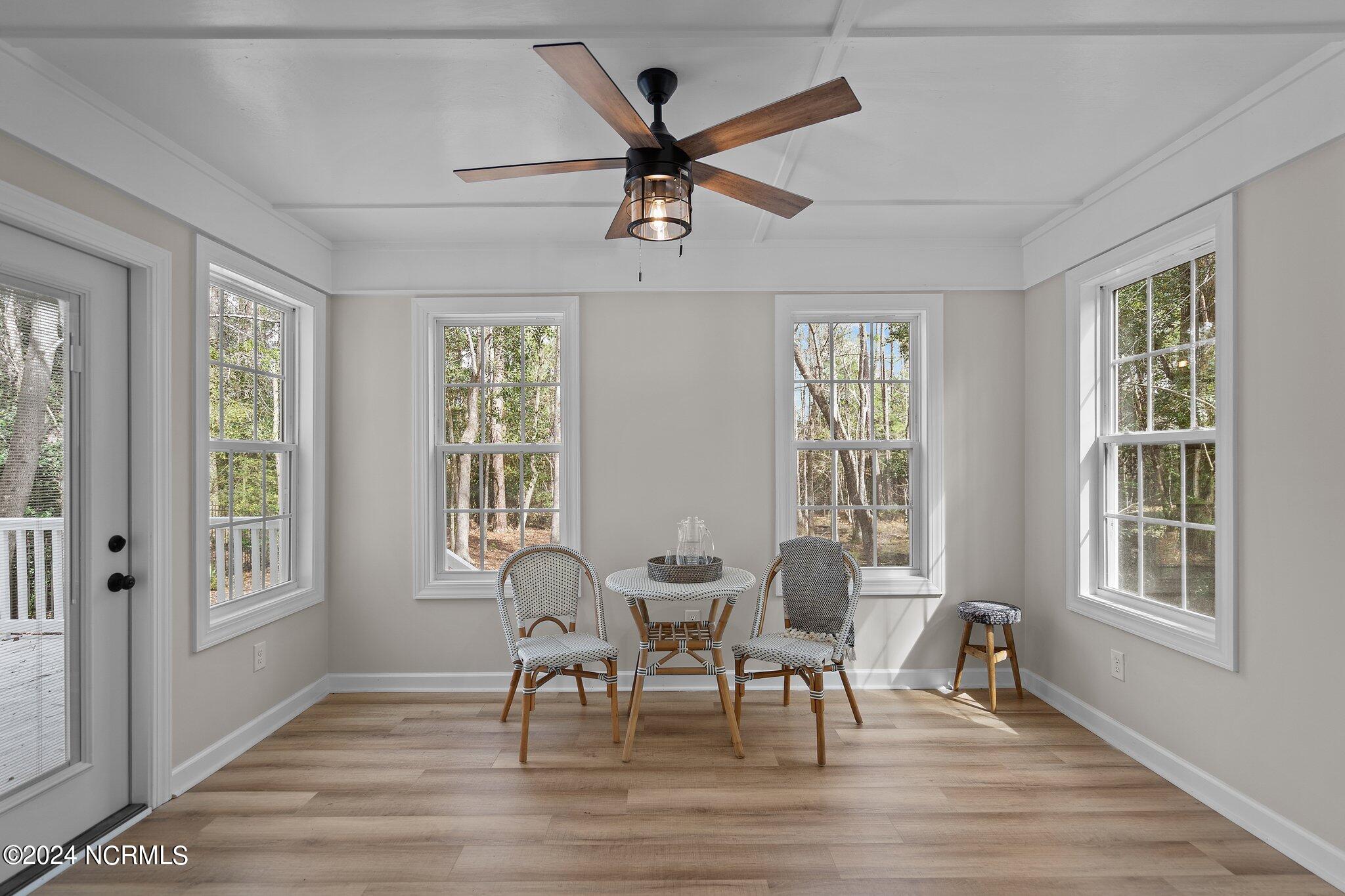 Furnished sunroom with a small table, woven chairs, and large windows overlooking trees.