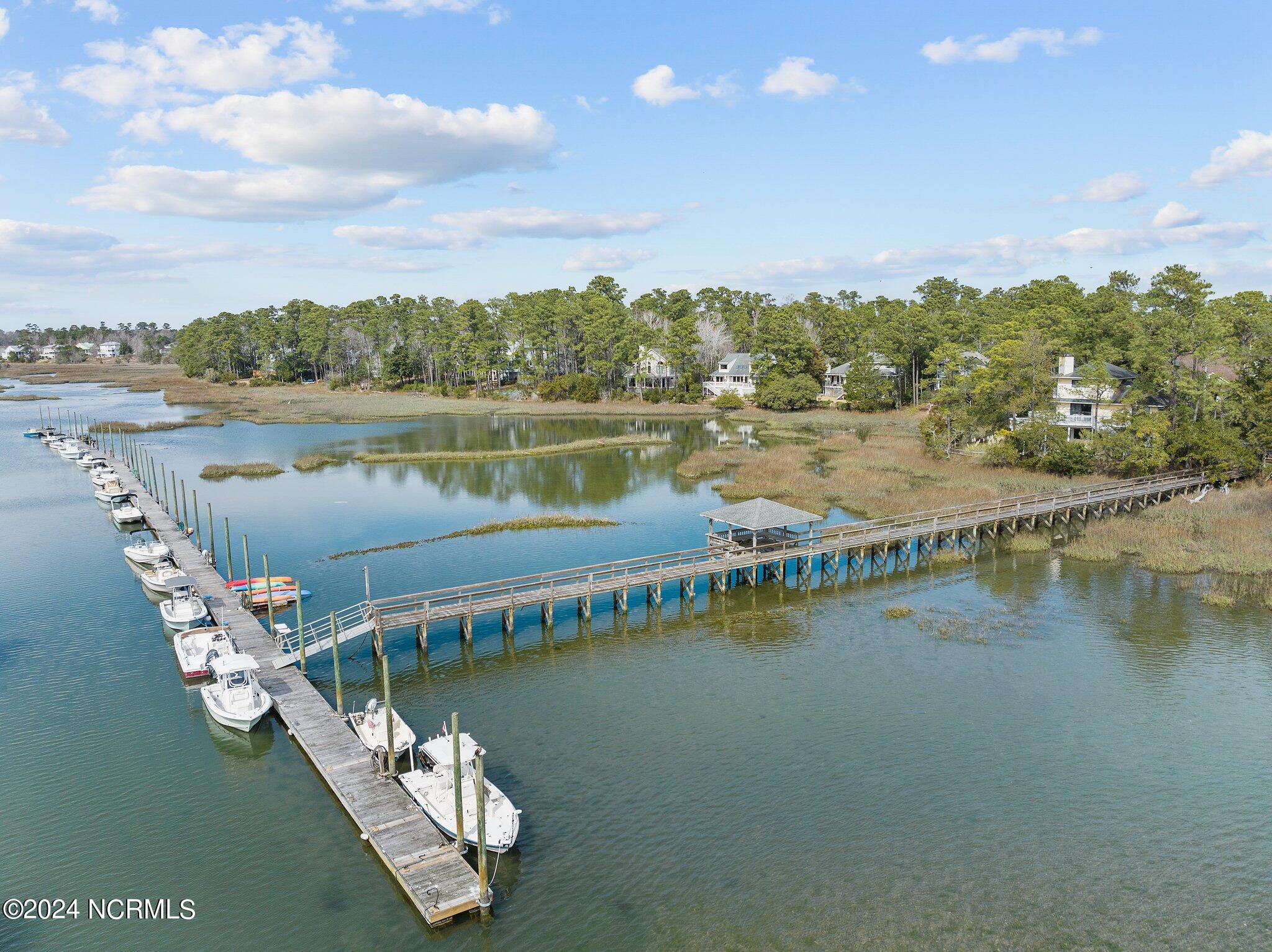 Aerial view of a creekside dock with moored boats, a covered pier, marshland, and homes along the wooded shore.