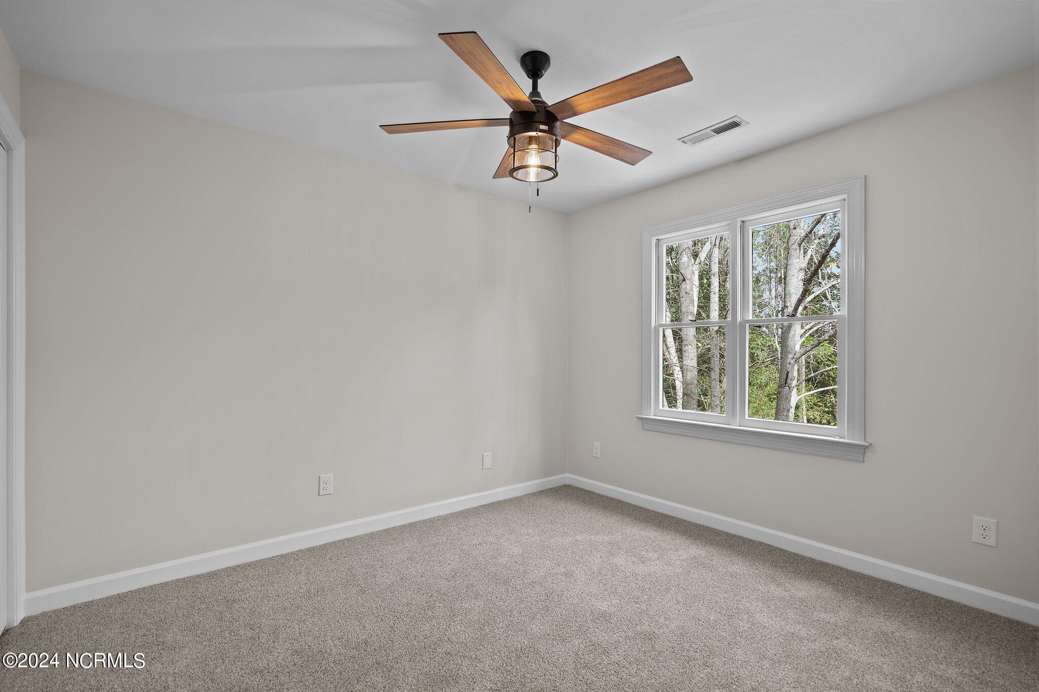 Empty carpeted bedroom with a wood-blade ceiling fan and double window overlooking trees.