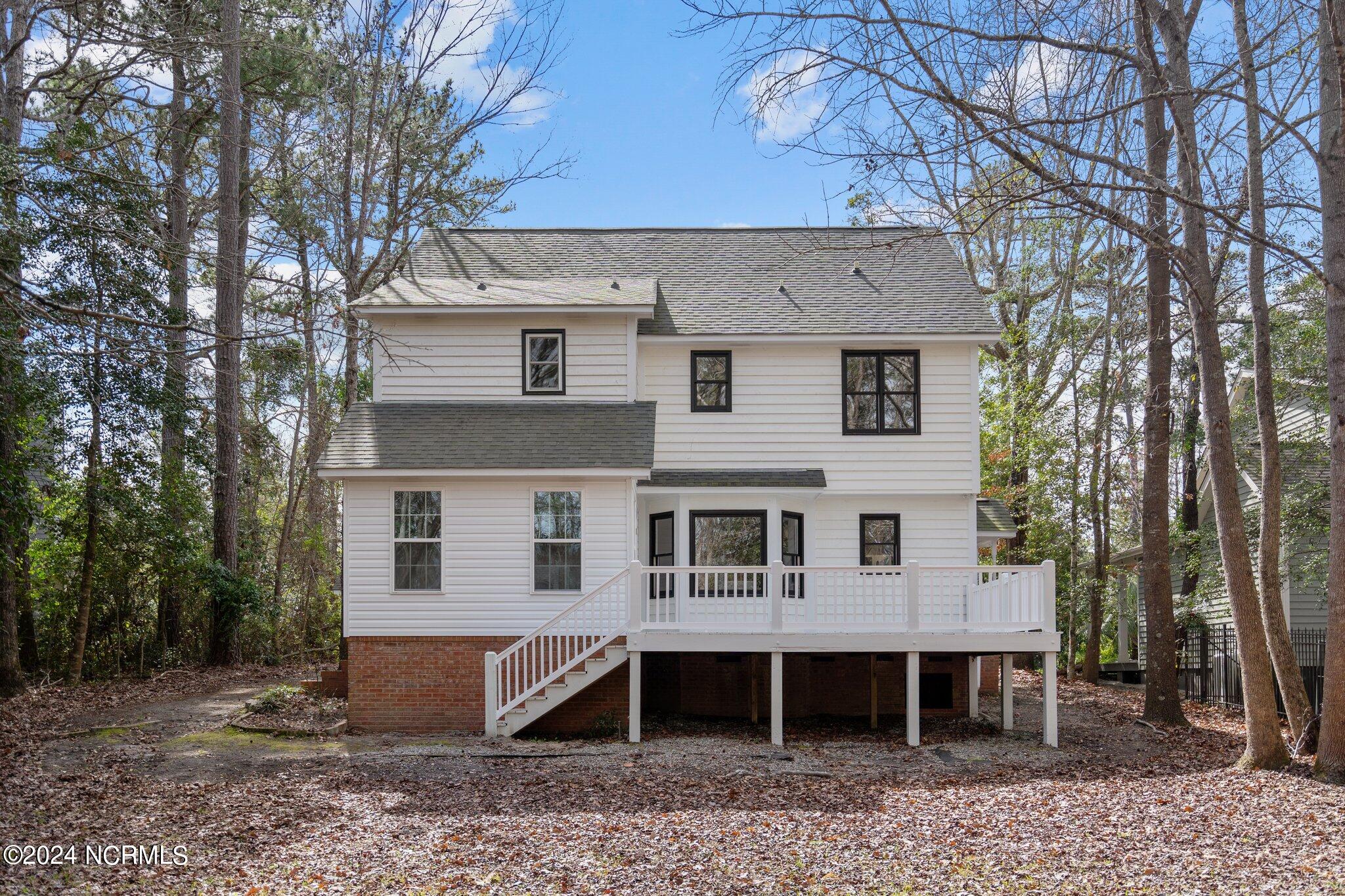 Rear of a two-story white house with a raised deck and stairs, surrounded by mature trees.