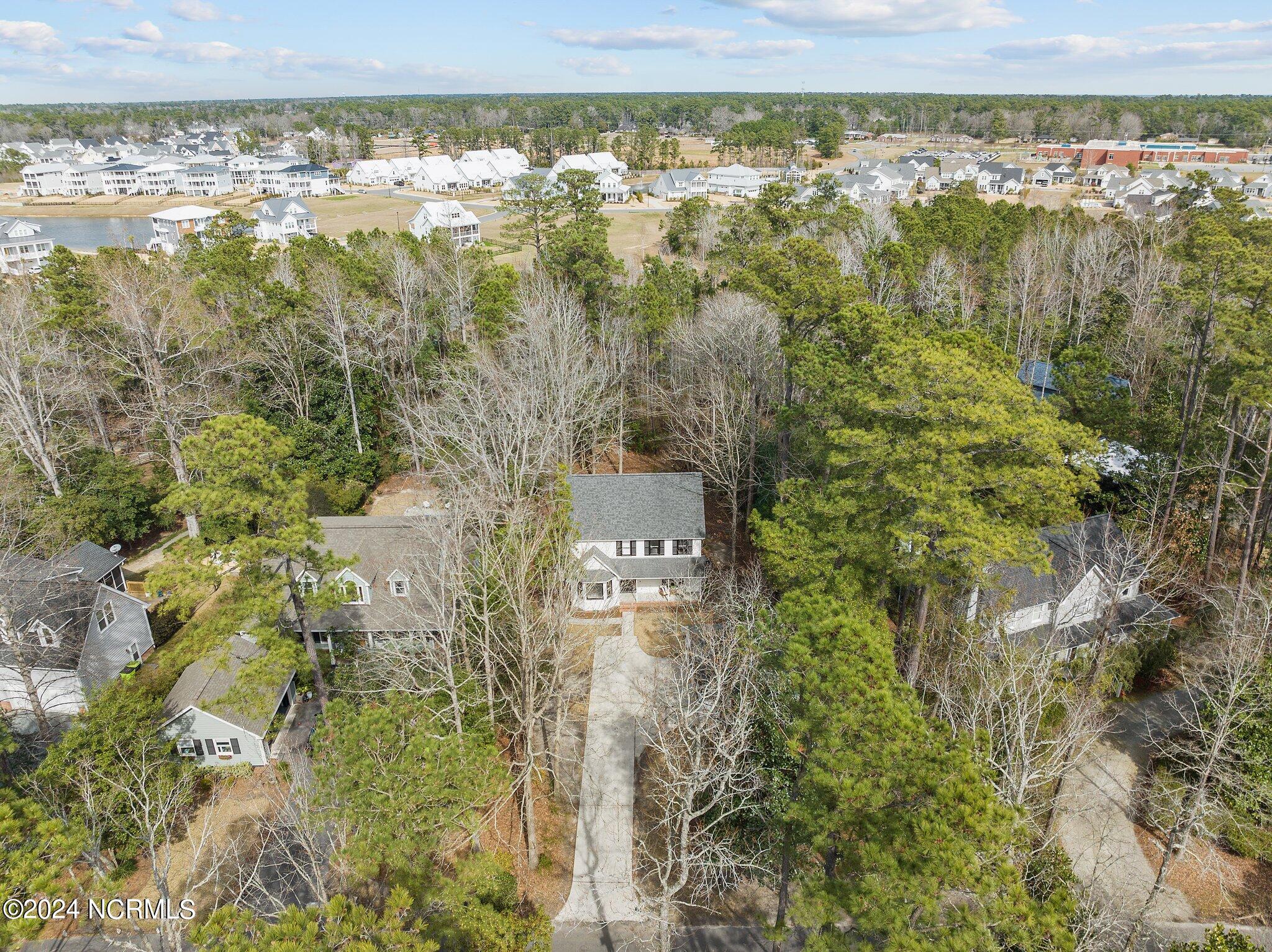 Aerial view of a wooded home with a long driveway and a residential neighborhood in the distance.