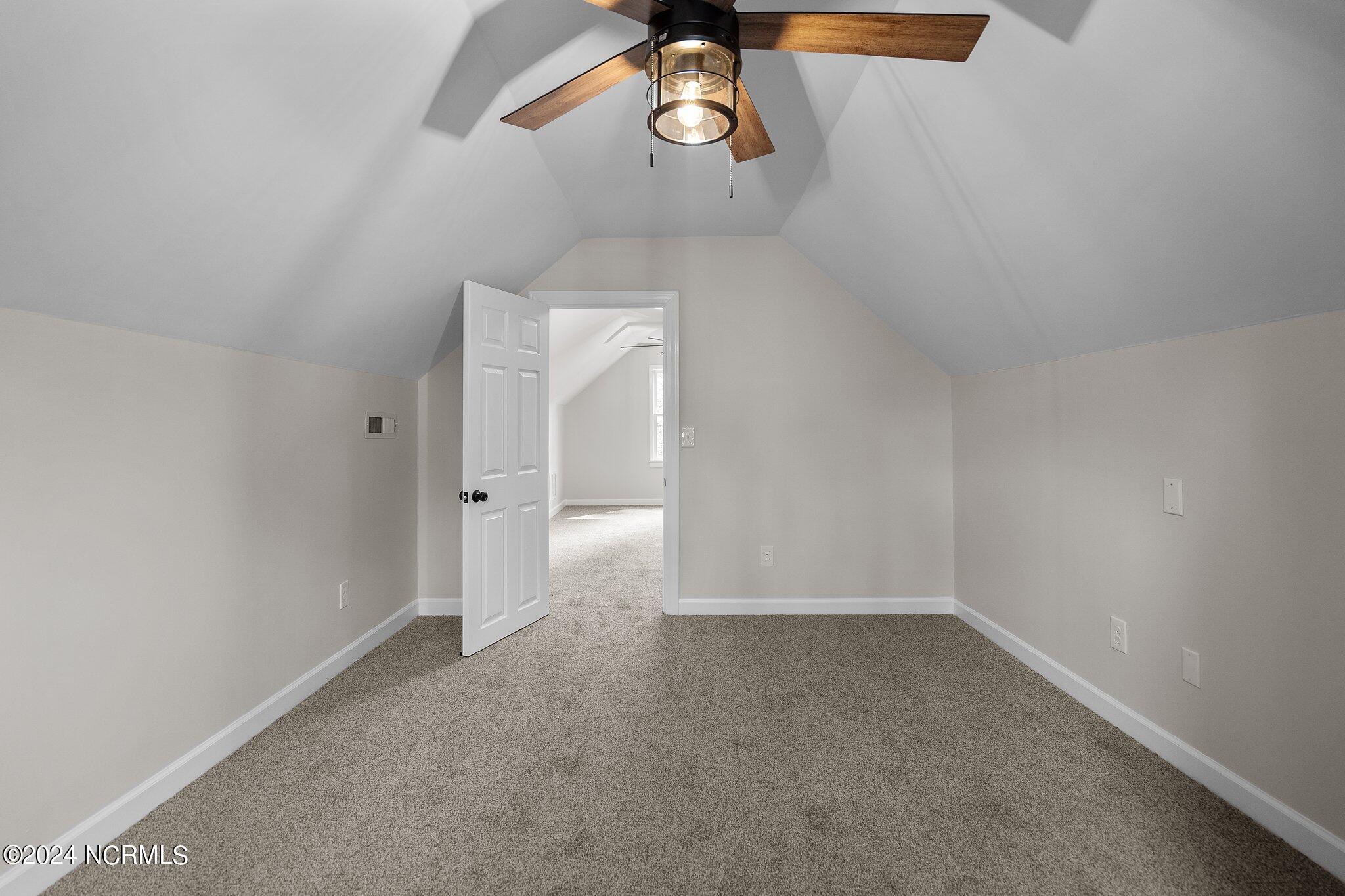 Empty carpeted bedroom with a vaulted ceiling and wood-blade ceiling fan, viewed toward an open doorway.
