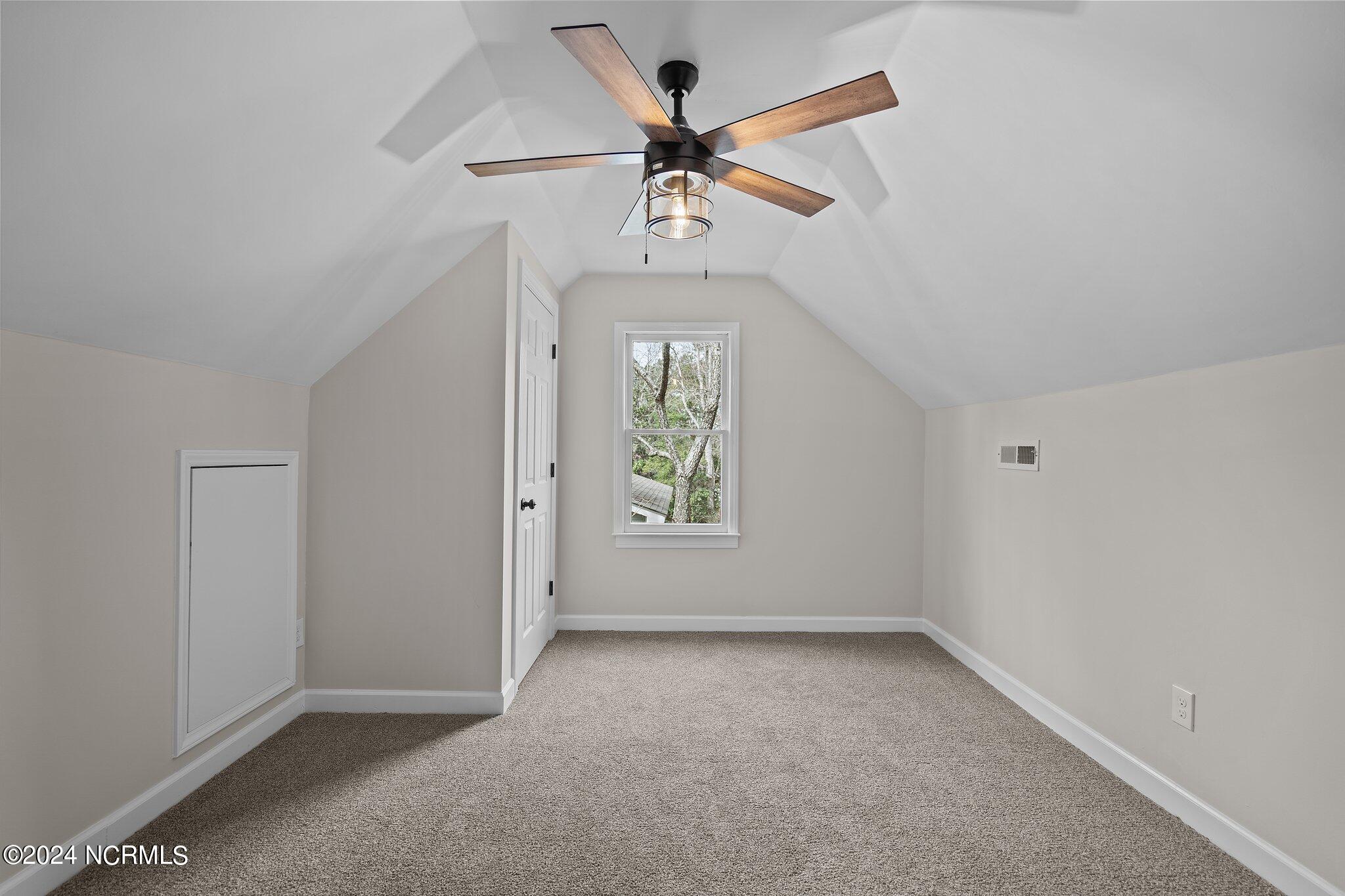 Empty carpeted room with a vaulted ceiling, wood-blade ceiling fan, and window.
