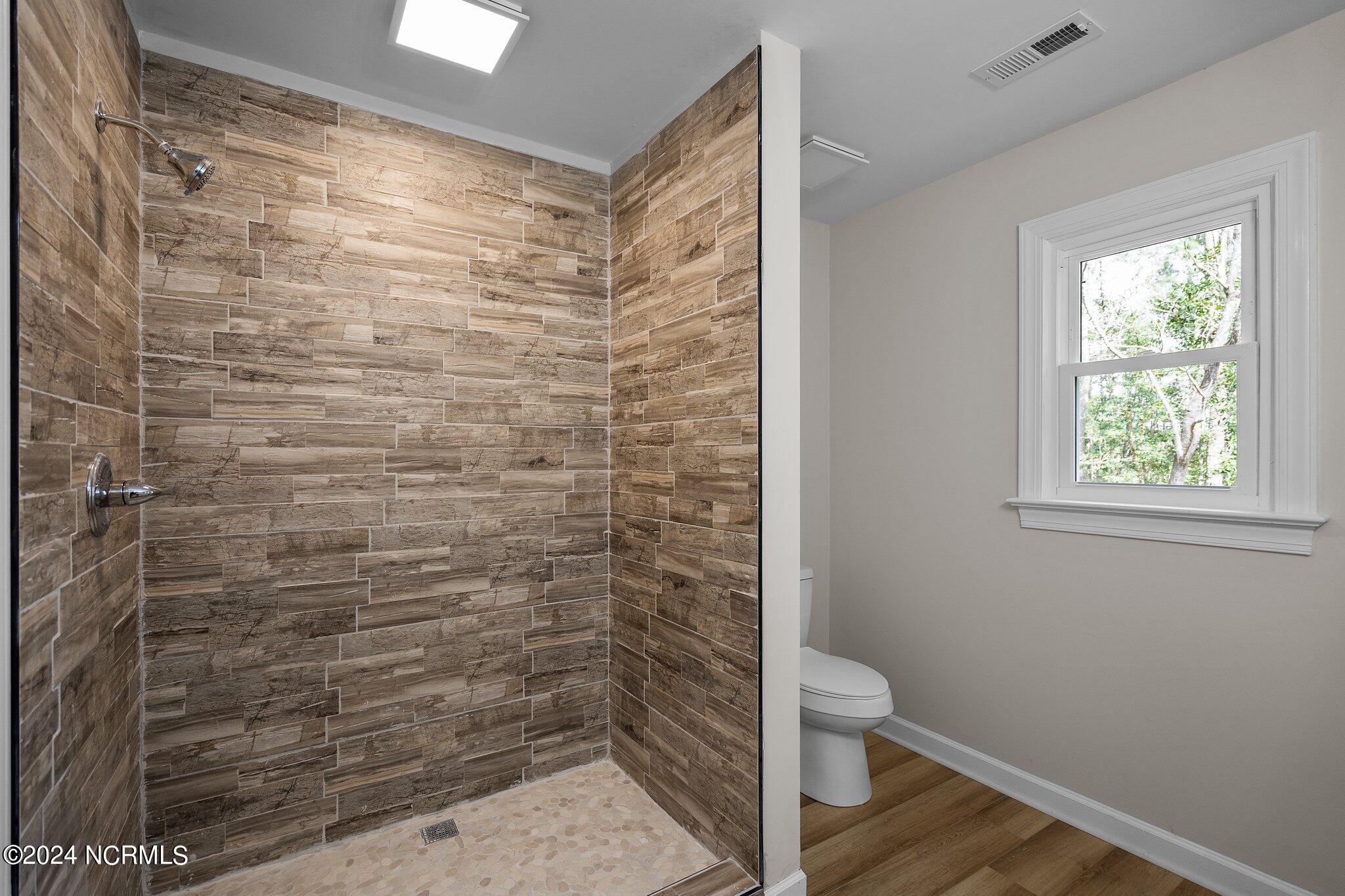 Bathroom with wood-look tiled walk-in shower, pebble floor, and toilet beside a window.