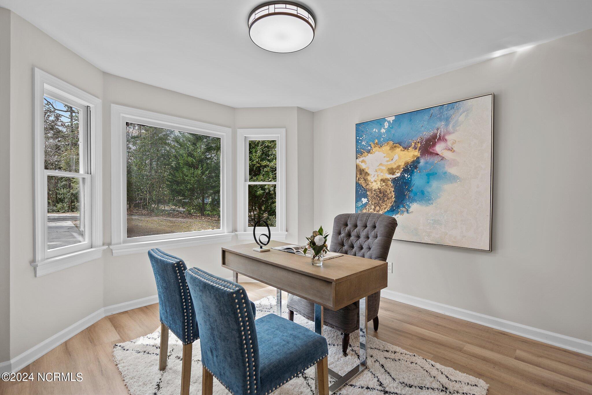 Furnished home office with a wooden desk, blue guest chairs, three windows, and a large abstract painting.