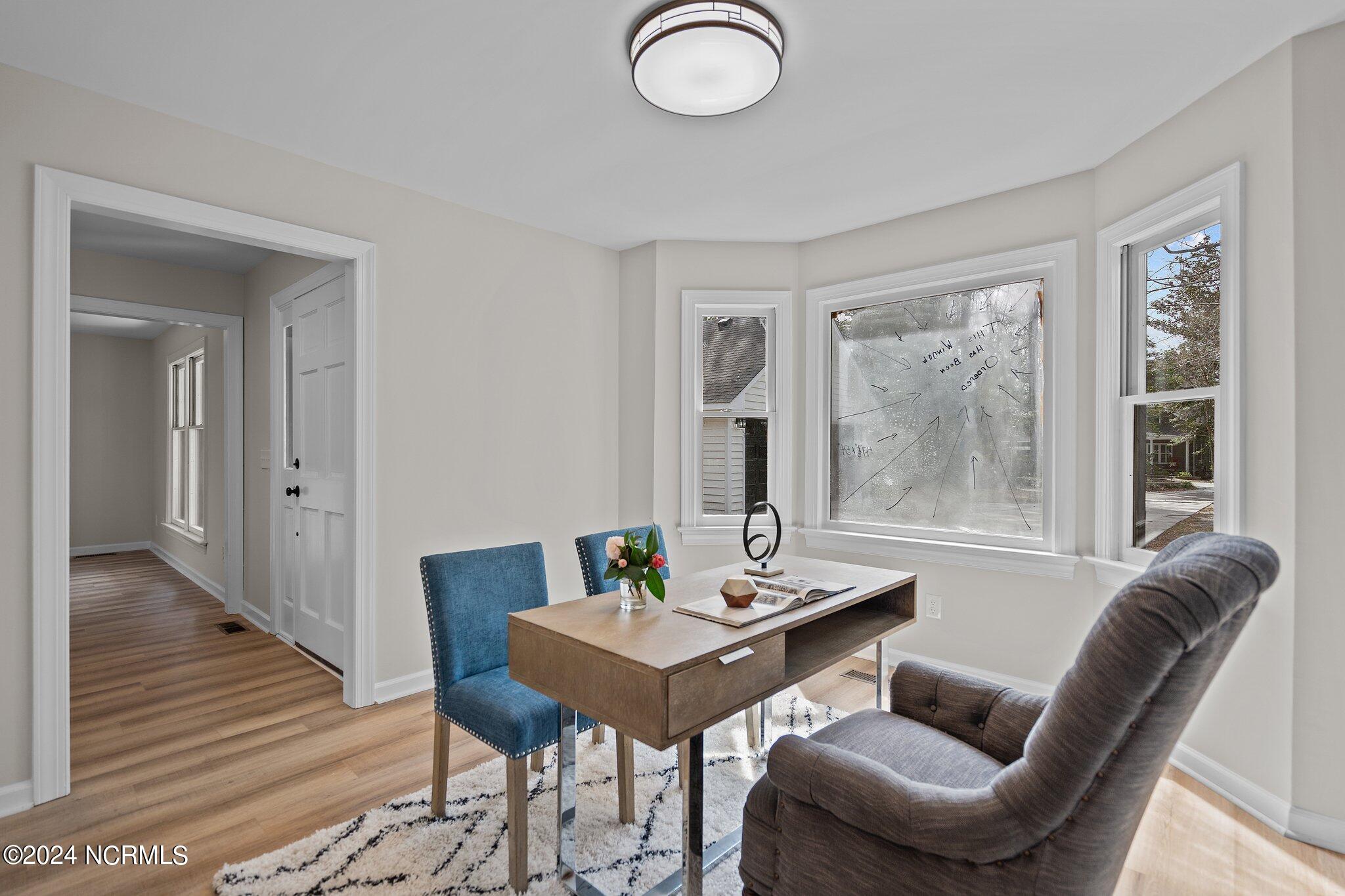 Staged home office with a wooden desk, blue chairs, gray armchair, and several windows.