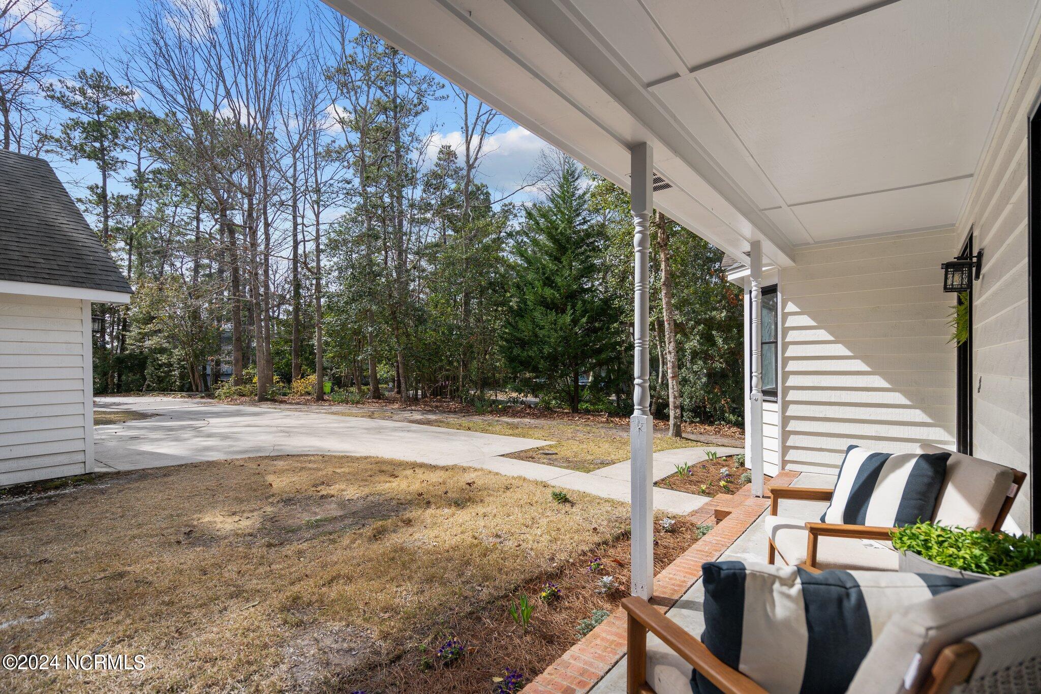 Covered porch with cushioned chairs overlooking a lawn, driveway, and mature trees.