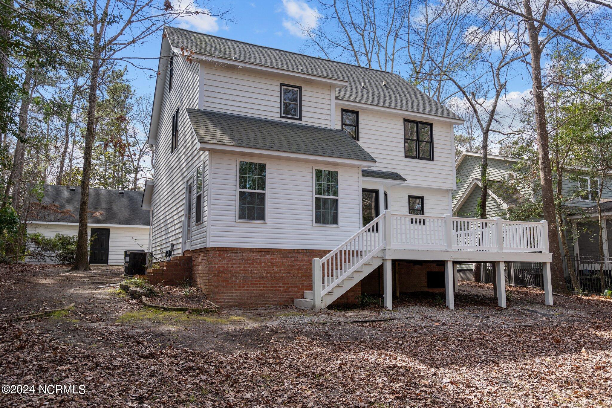 White two-story house with a raised deck and stairs in a wooded, leaf-covered yard.