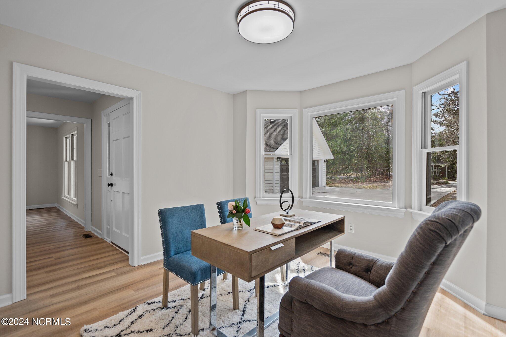 Staged home office with a wooden desk, blue chairs, and large windows overlooking trees.
