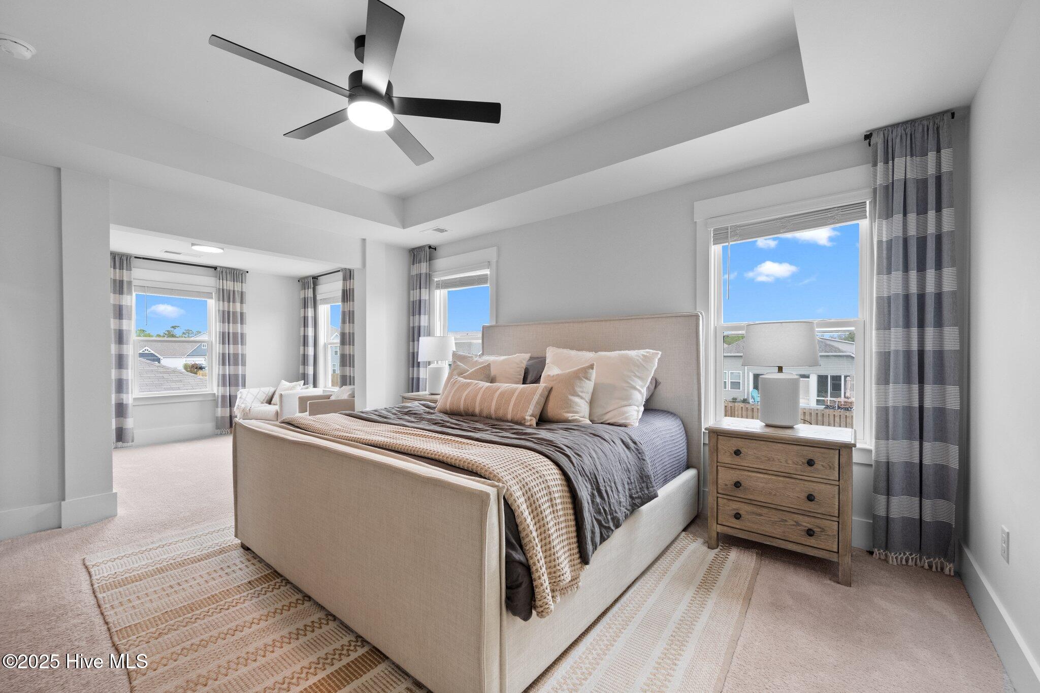 Staged bedroom with an upholstered bed, tray ceiling, and a small sitting area by three windows.