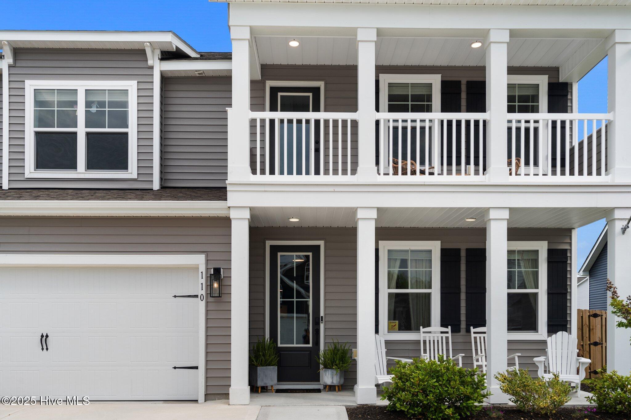 Two-story gray home with a garage, covered upper porch, and white chairs on the lower porch.
