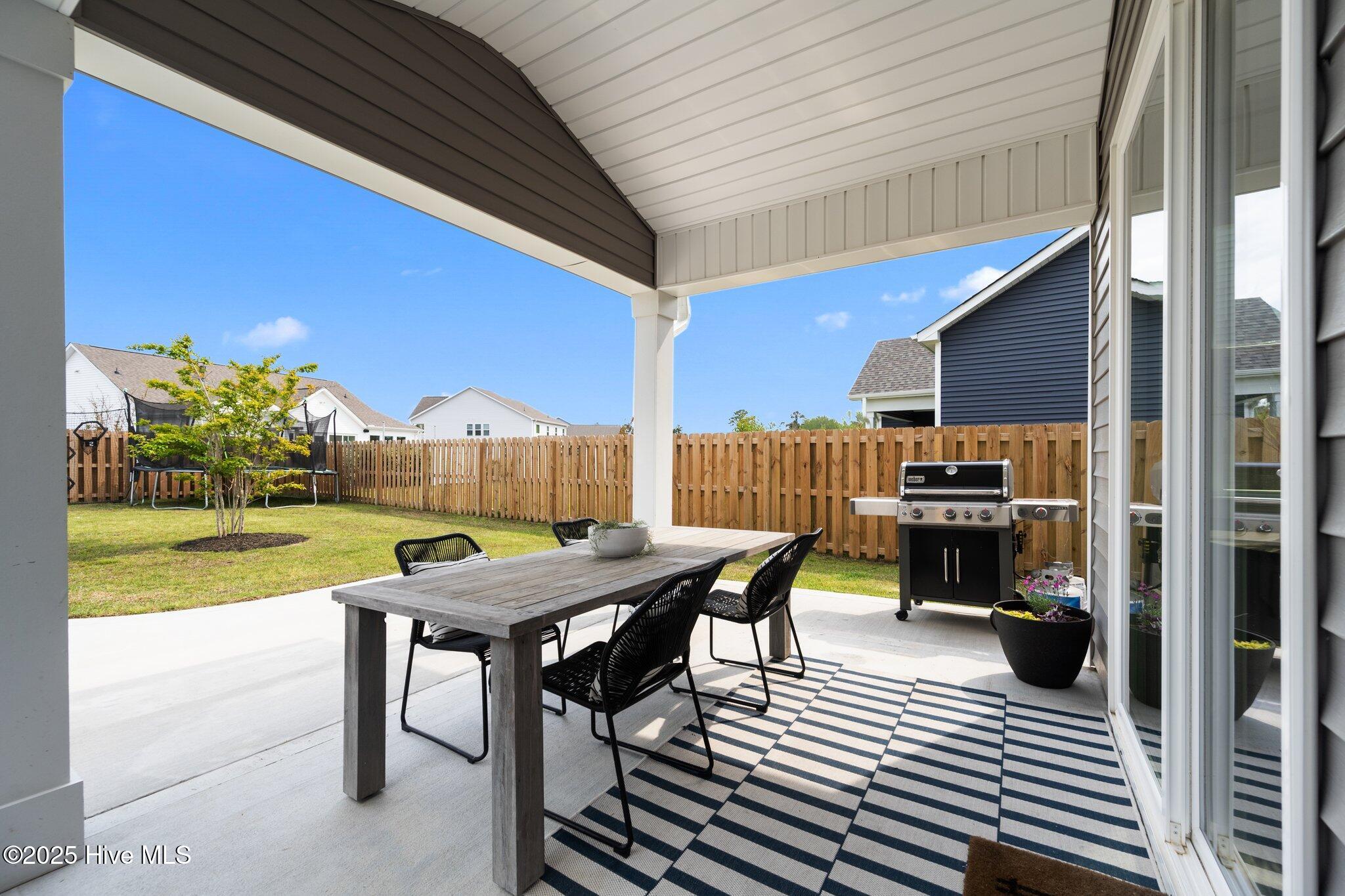 Covered patio with outdoor dining set, gas grill, striped rug, and fenced lawn.