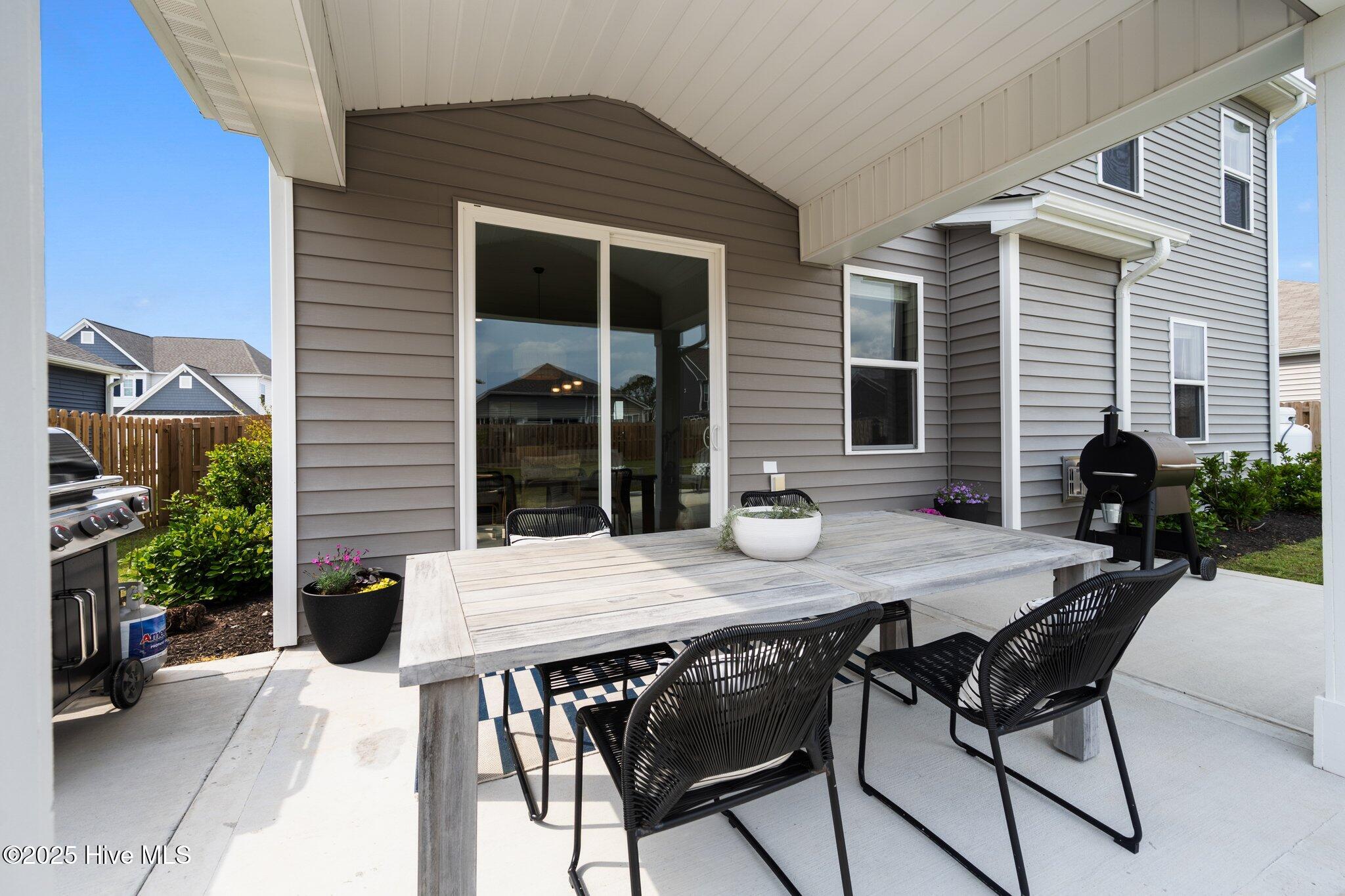 Covered patio with a large dining table, black chairs, and grills beside the gray-sided home.