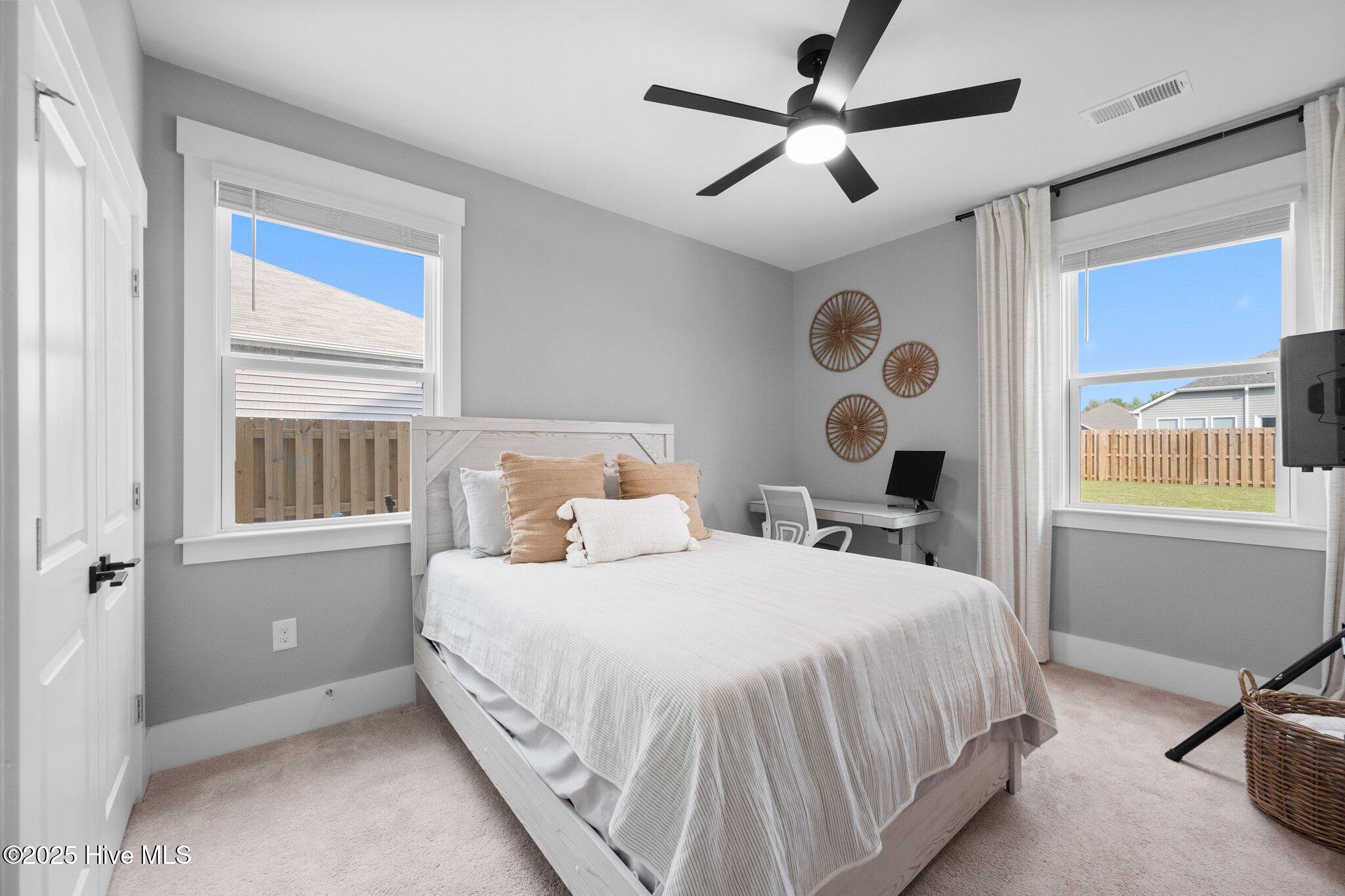 Staged bedroom with a white bed, black ceiling fan, desk, and windows overlooking a fenced yard.