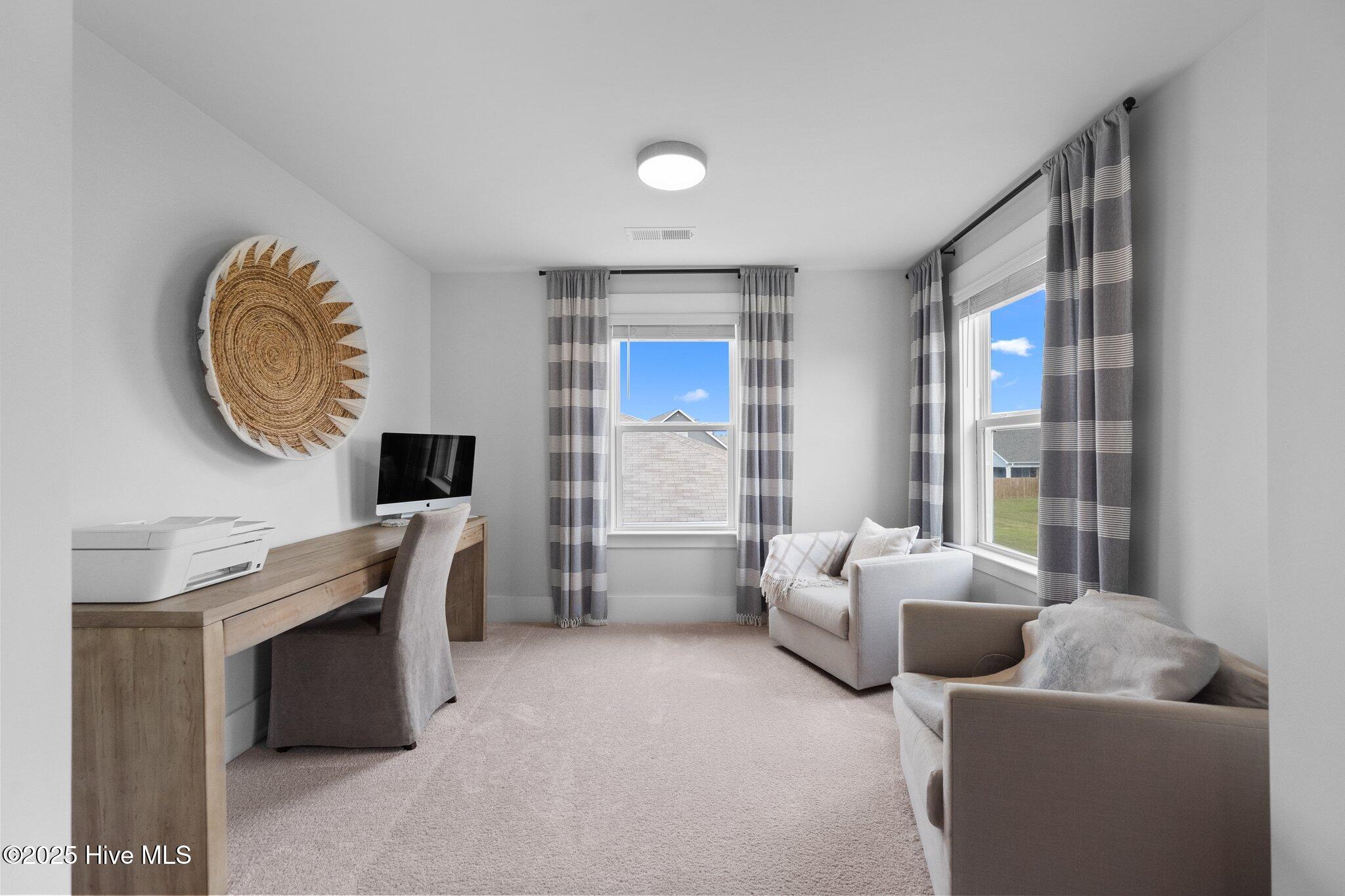 Furnished home office with a wooden desk, two upholstered chairs, and bright windows with striped curtains.