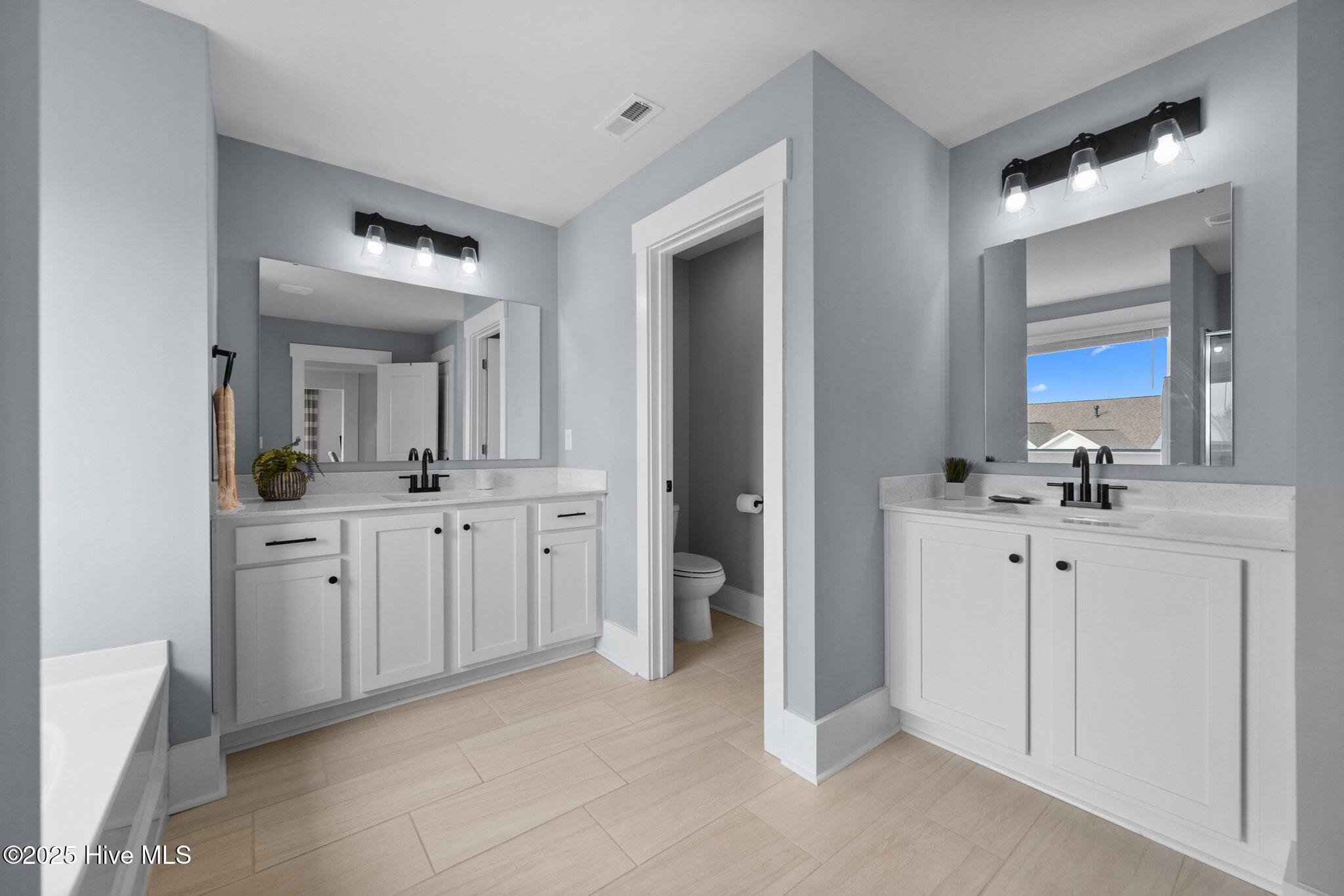 Bathroom with dual white vanities, black faucets, large mirrors, and a separate toilet alcove.