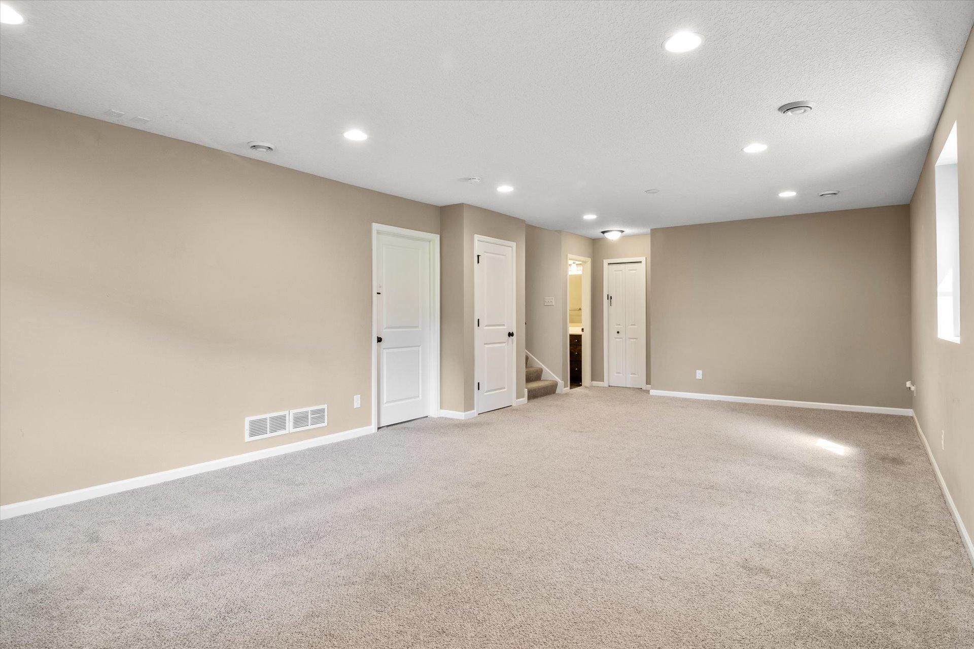 Empty carpeted room with beige walls, recessed lights, several white doors, and stairs at the back.
