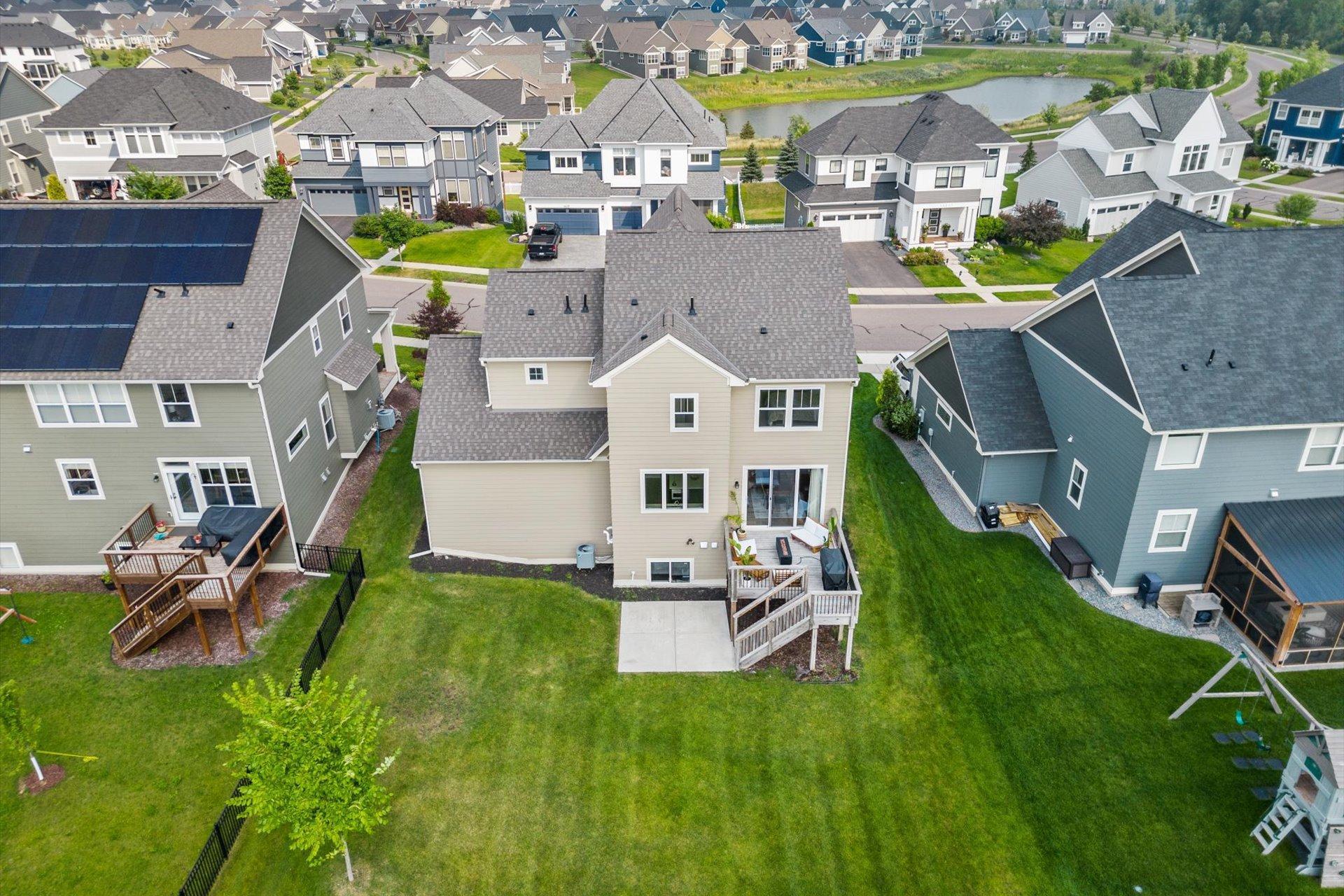 Aerial view of a beige two-story home’s backyard, deck, patio, and lawn, with a pond beyond neighboring homes.