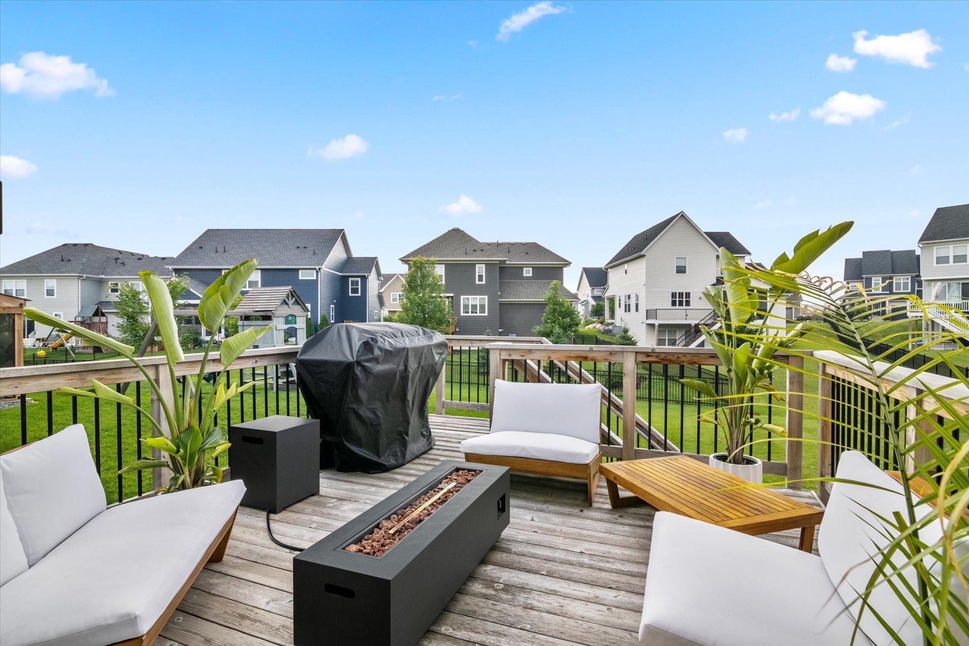 Furnished backyard deck with white lounge chairs, a covered grill, and a fire table overlooking neighboring homes.