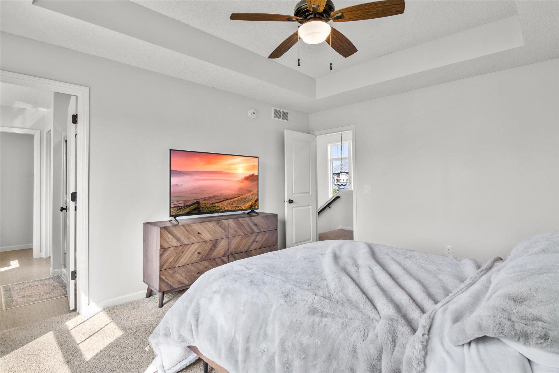 Bright bedroom with a white bed, wood dresser and television beneath a tray ceiling.