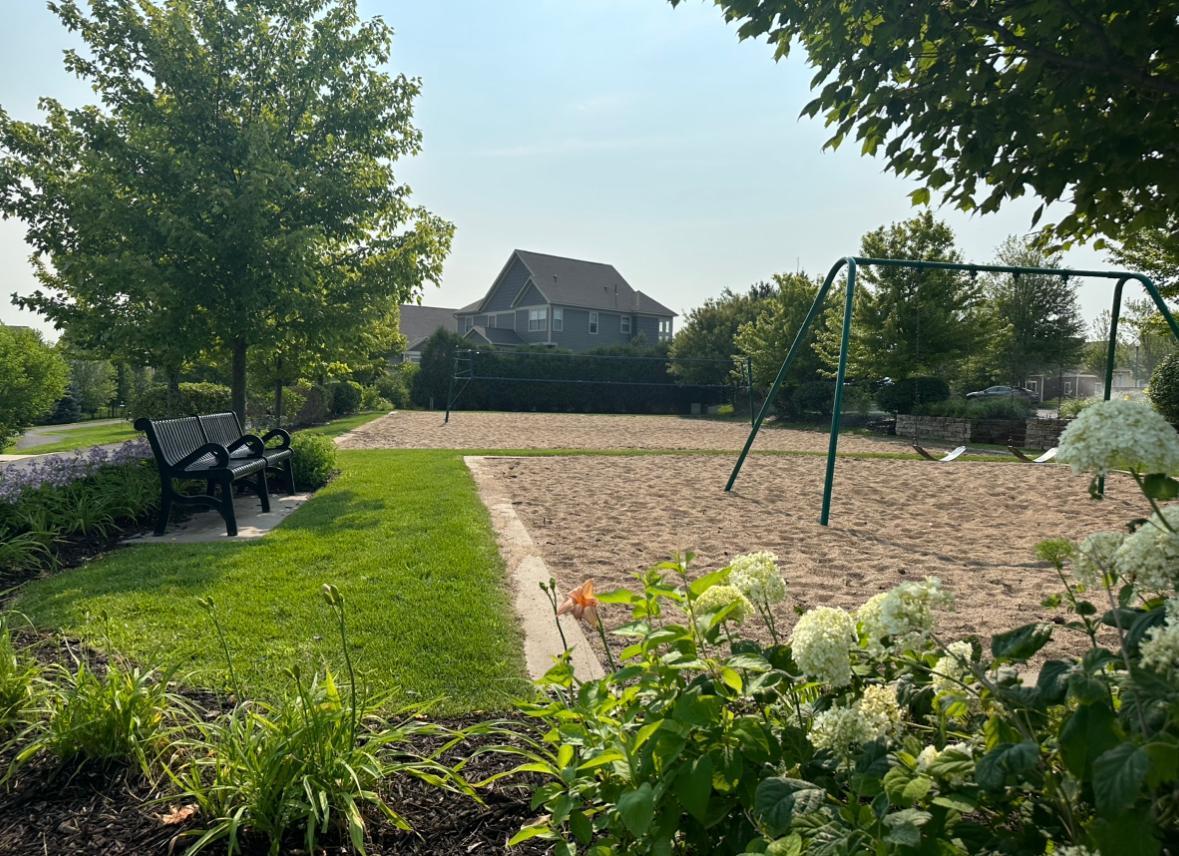 Landscaped playground with sandy play area, swings, and bench beneath mature trees.