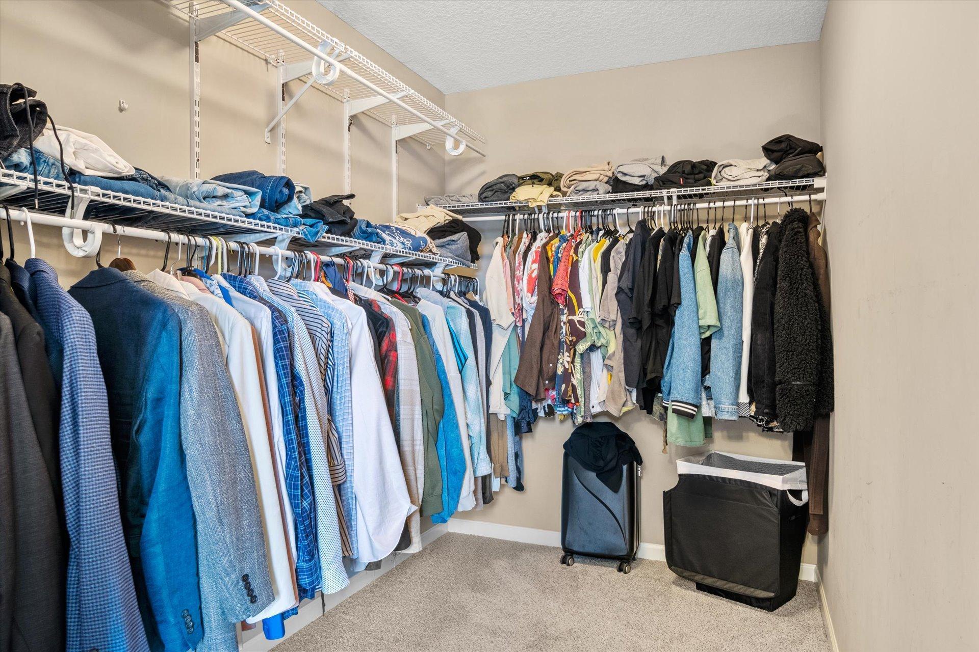 Walk-in closet with wire shelving filled with clothes and a suitcase on the carpet.