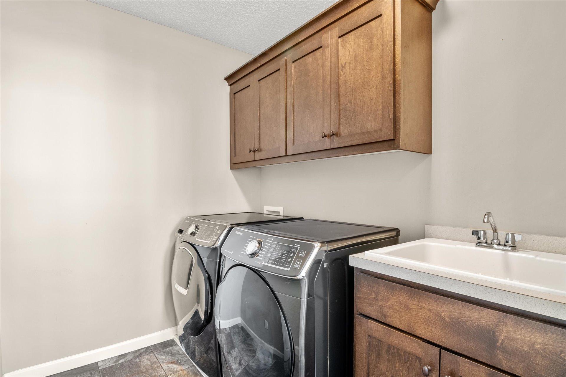 Laundry room with front-loading washer and dryer beneath wood cabinets, beside a utility sink.