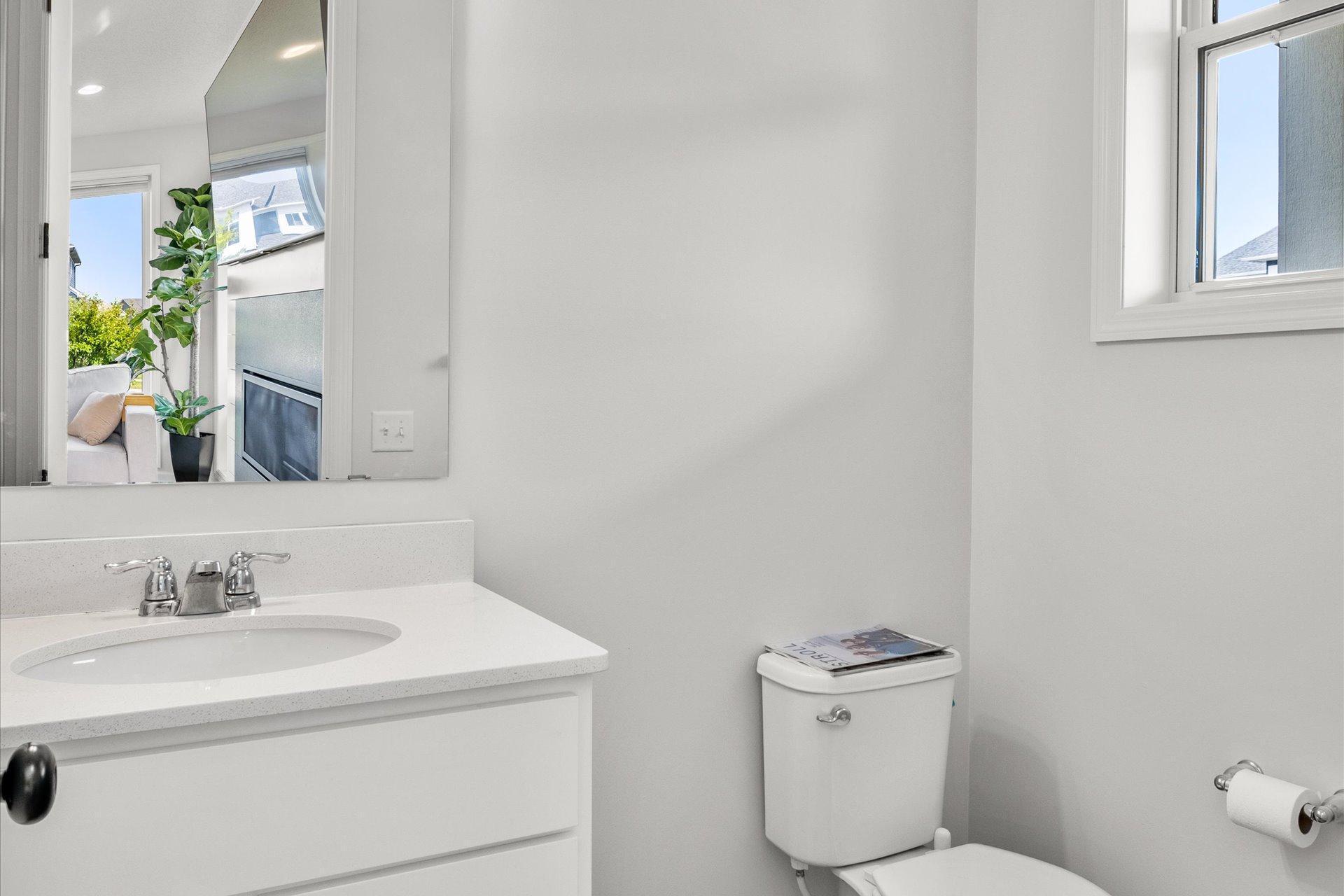 Bright bathroom with a white vanity and toilet; the mirror reflects a living area and fireplace.