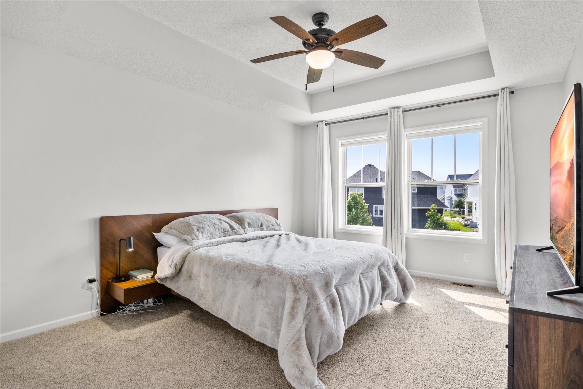 Furnished bedroom with a large bed, ceiling fan, and two windows overlooking neighboring homes.